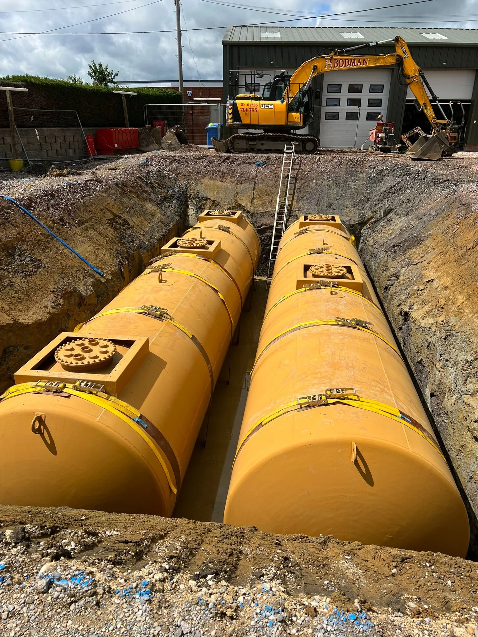 Two large yellow underground storage tanks installed in a deep excavation at a construction site, with an excavator parked on the edge and a ladder leading up to the surface.
