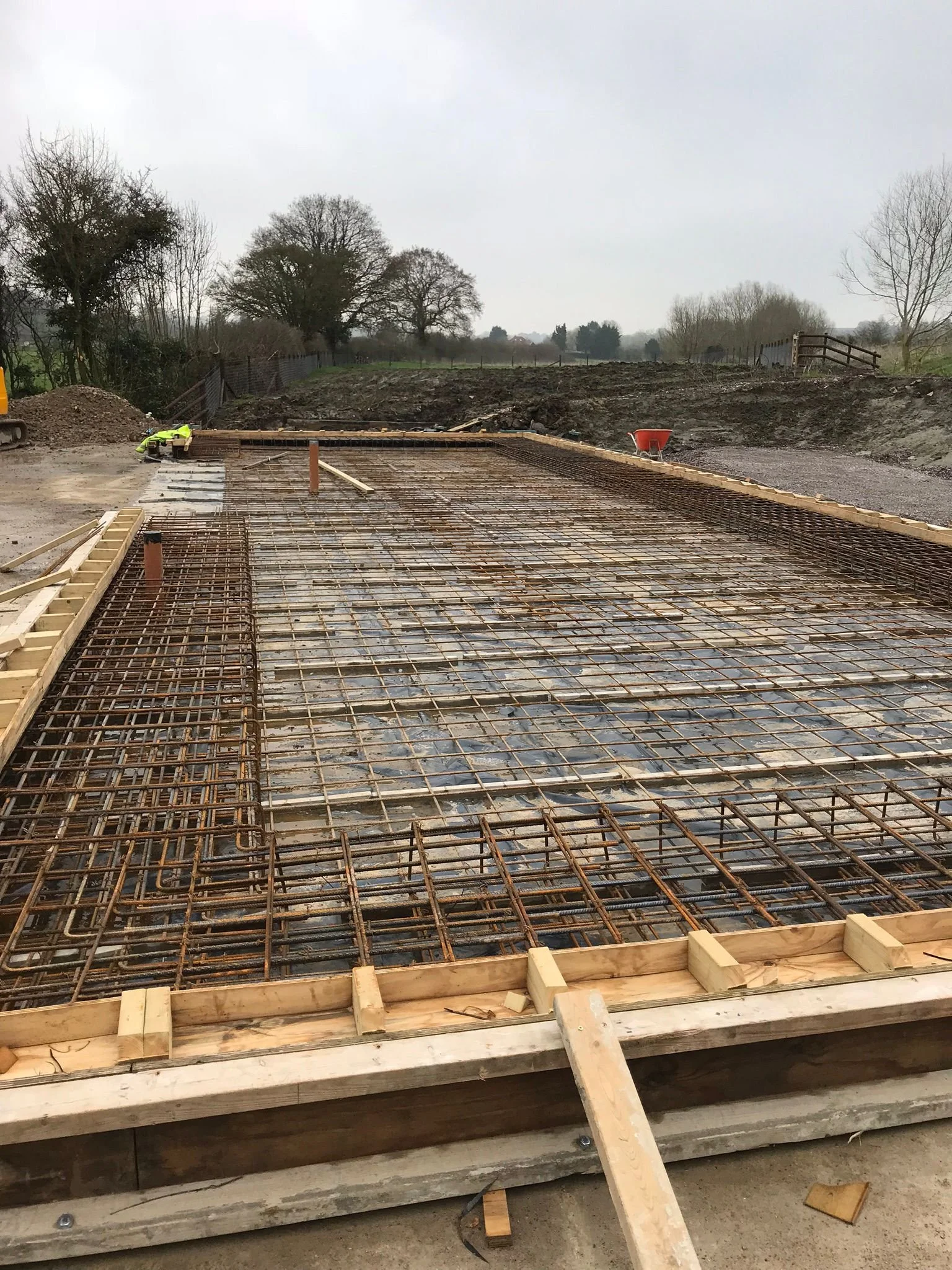 Construction site with rebar grid prepared for concrete pouring, wooden formwork edges, and surrounding dirt landscape.