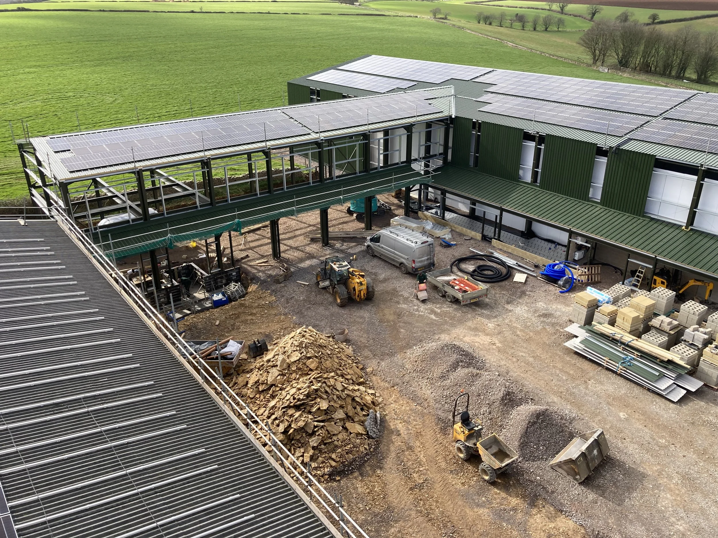 Construction site of a building with solar panels on the roof, surrounded by green fields, showing construction materials, machinery, and partially completed structures.