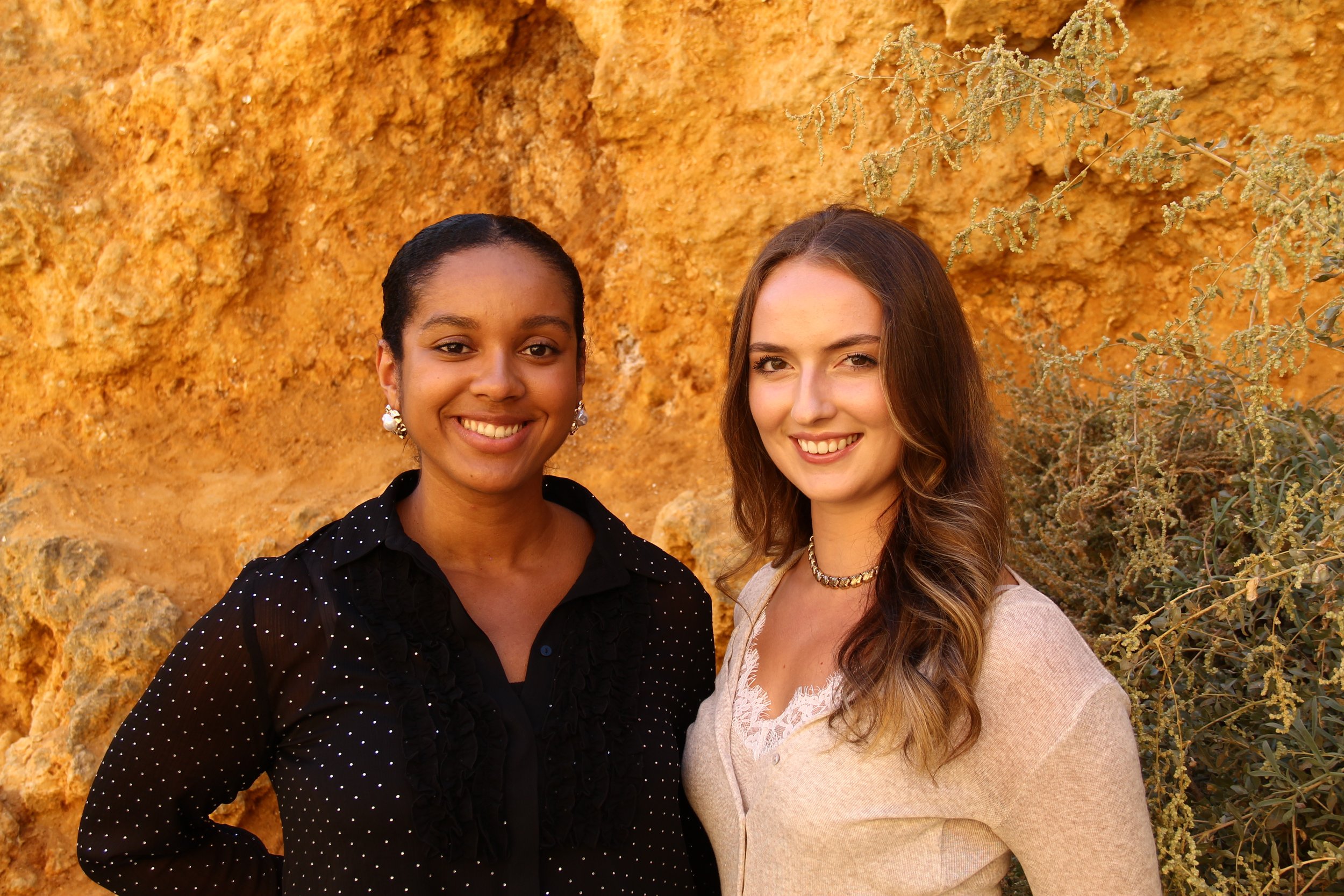 Two women smiling outdoors against a yellowish rock background. The woman on the left has dark skin, black hair pulled back, and is wearing a black polka-dot blouse with earrings. The woman on the right has light skin, long wavy brown hair, and is wearing a beige sweater with a necklace.