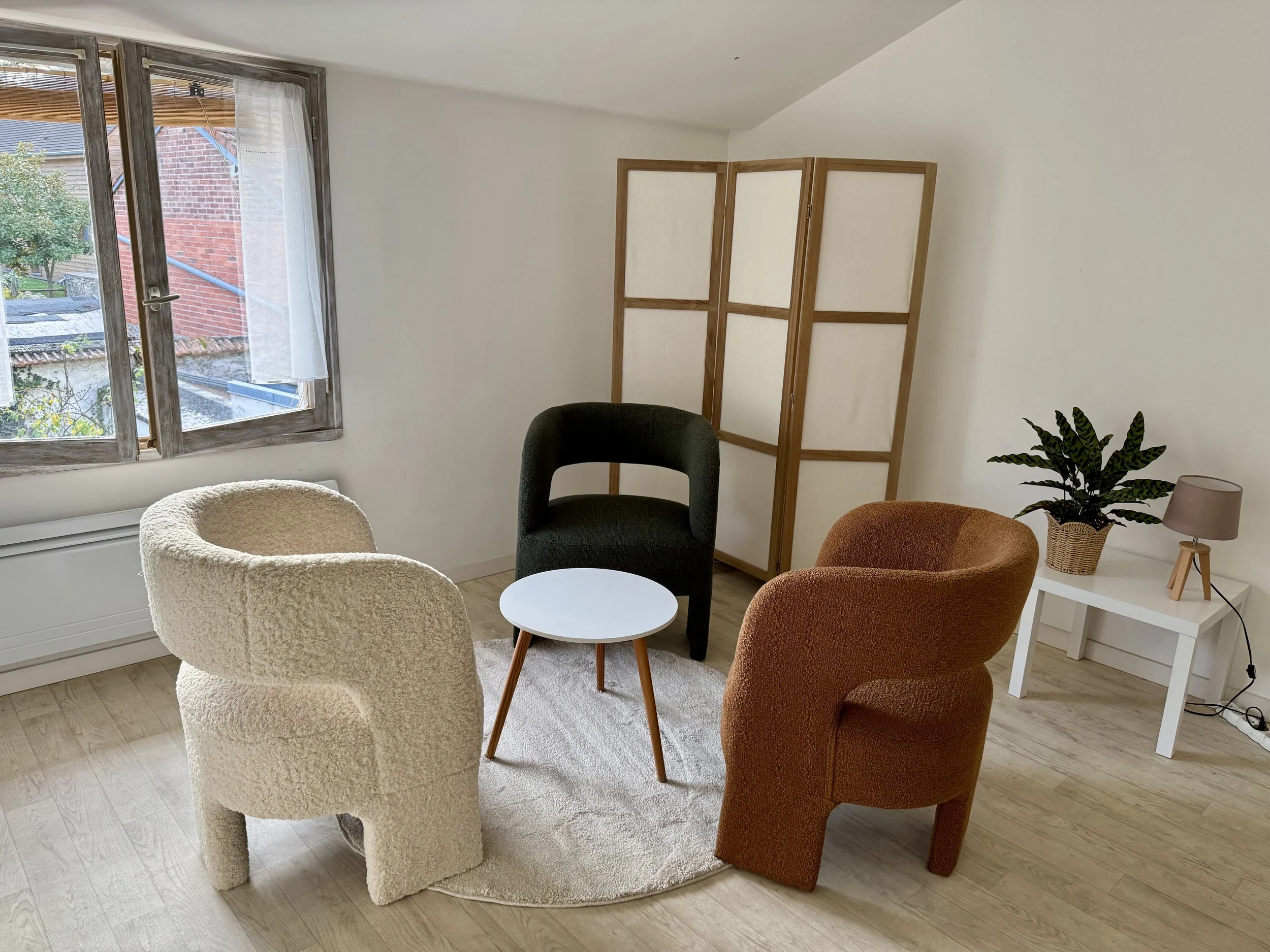 Salon avec trois fauteuils (beige, noir, orange) autour d'une petite table ronde blanche, à côté d'une fenêtre. Consultation évasion et mur blanc avec panneau en bois. Plante verte sur une table blanche avec une petite lampe. Sol en bois et tapis beige.