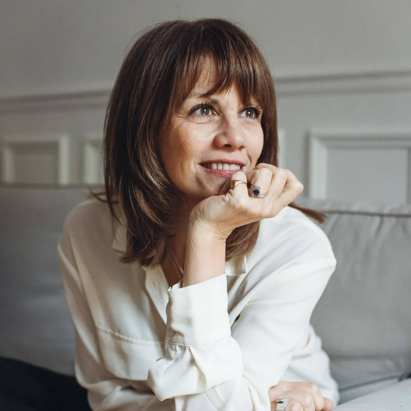 Une femme aux cheveux bruns, souriante, portant une chemise blanche, assise dans un salon, posant avec sa main sous son menton.