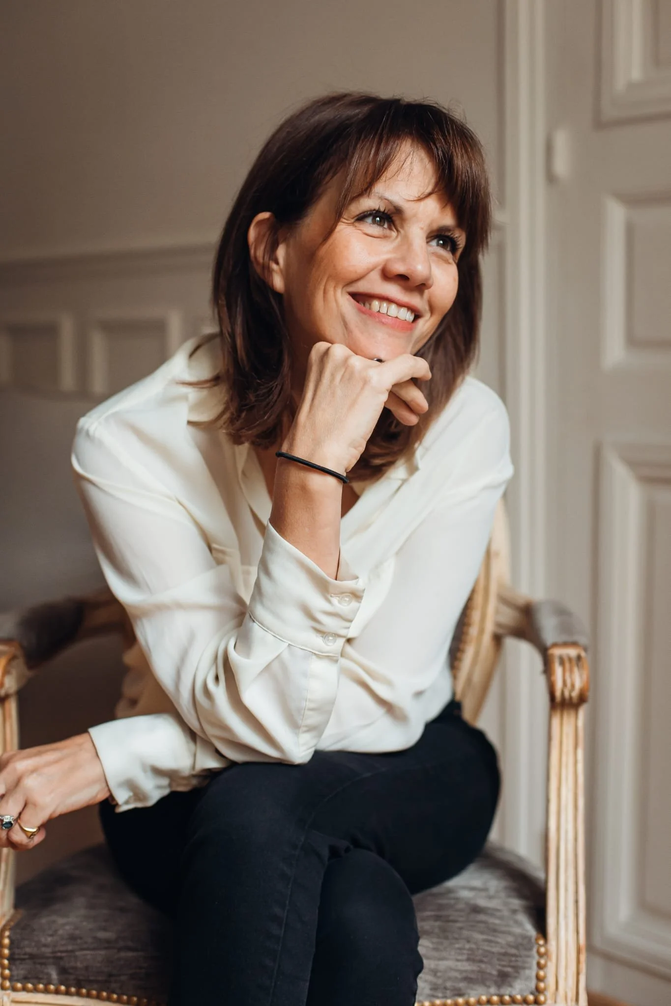 Une femme souriante assise dans un fauteuil en bois, portant une chemise blanche et des pantalons noirs, à l'intérieur d'une pièce avec des murs beiges et une porte blanche.