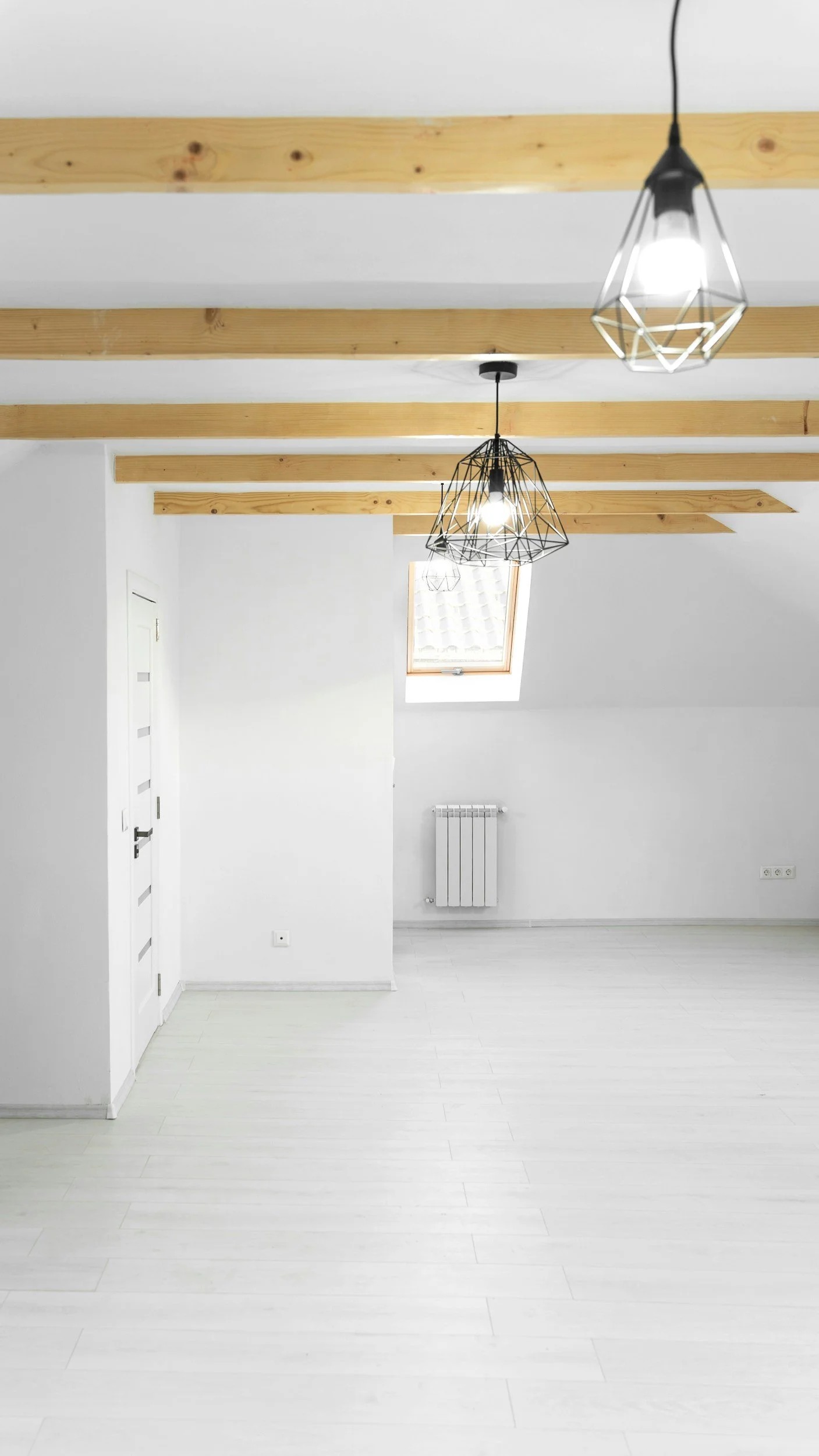 Empty room with white walls, light-colored wooden flooring, exposed wooden ceiling beams, a small skylight window, and modern pendant lights.