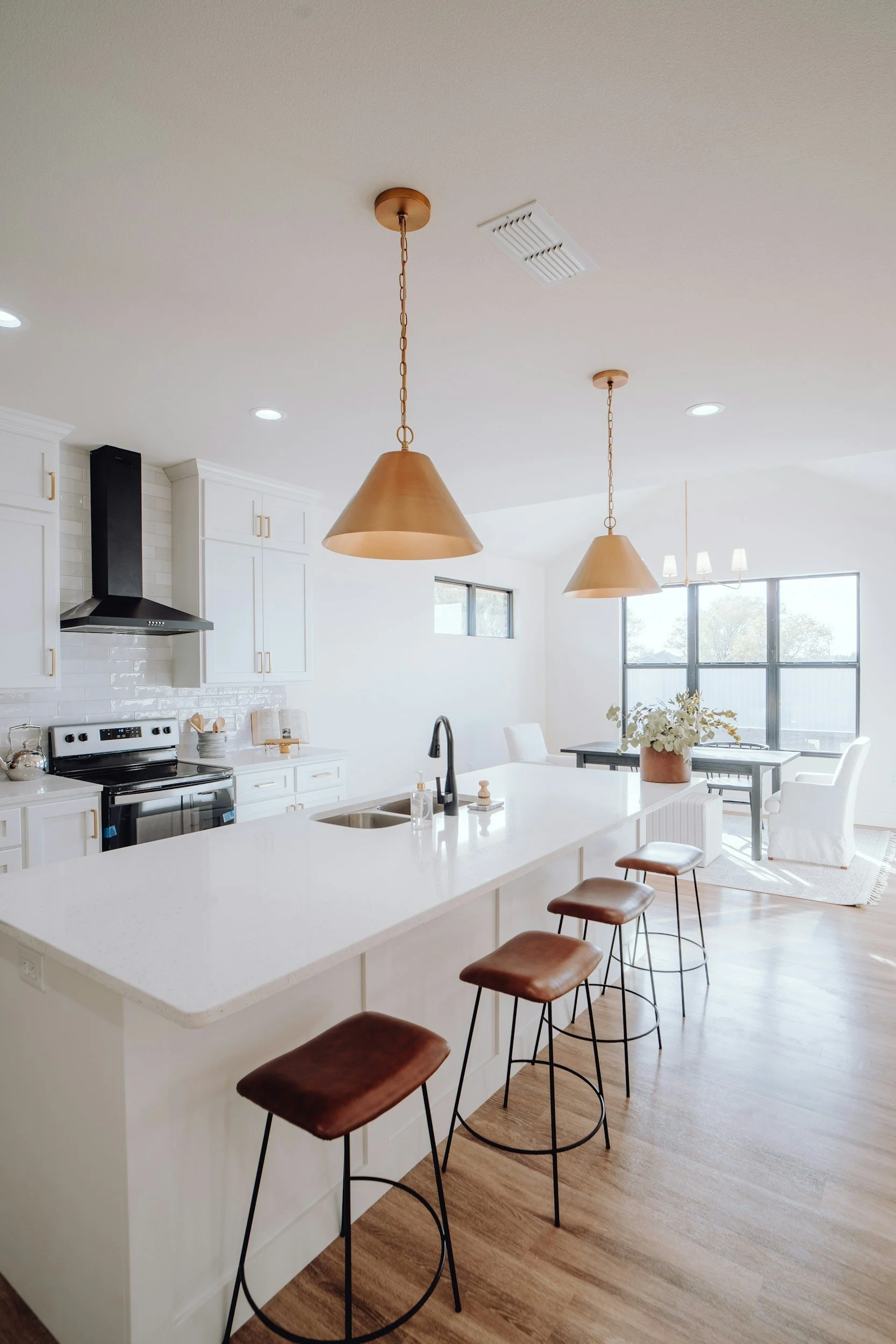 Bright, modern kitchen with white cabinets and island, three brown bar stools, black faucet, and at the back a dining area with large window, white chairs, and a vase with flowers.
