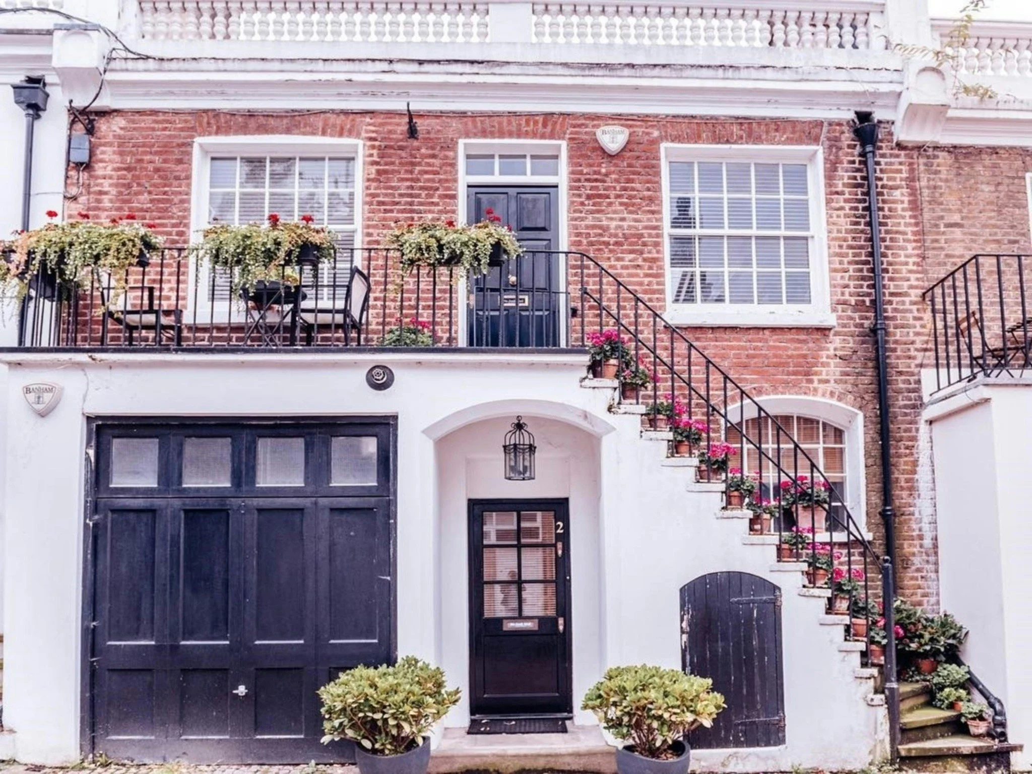 Front view of a brick townhouse with a black garage door, a black front door, large windows with white trim, and a small balcony with potted plants and flowers.