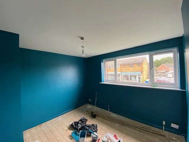 Empty room with teal walls, a large window, and a ceiling light fixture. Construction tools and supplies are on the floor.
