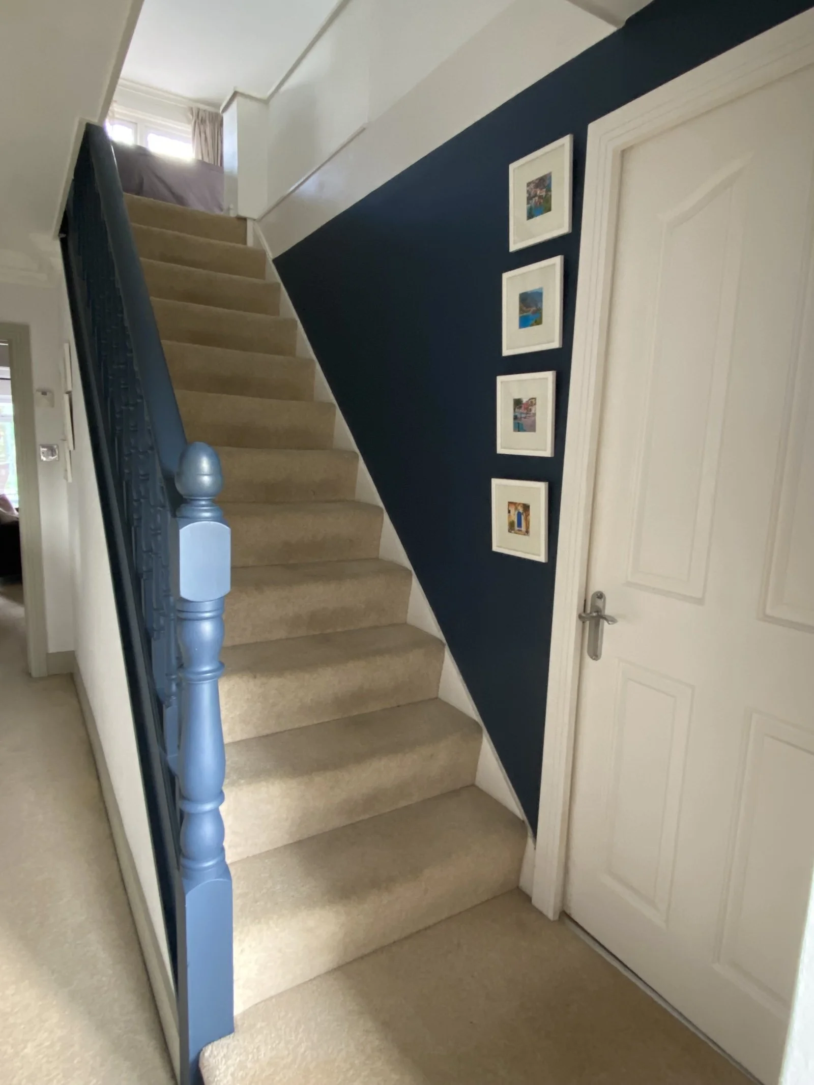 Interior view of a beige-carpeted staircase with a blue wall on one side, four framed pictures on the wall, and a white door at the bottom.