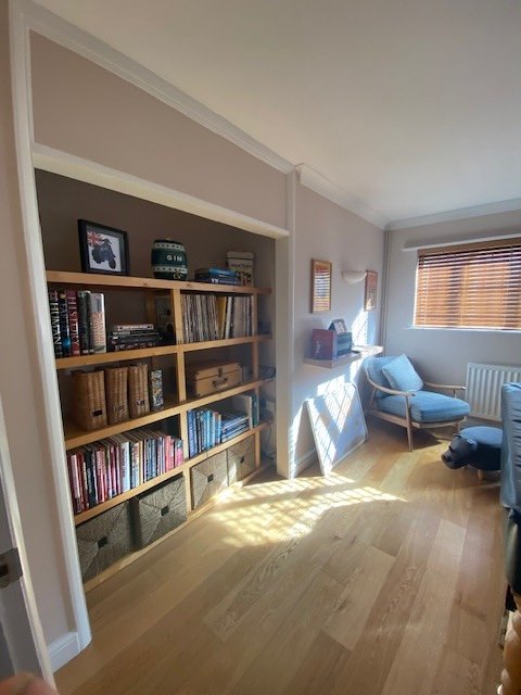Living room corner with a wooden bookshelf filled with books and decorative items, a blue armchair, a small side table, and a window with wooden blinds letting in sunlight.