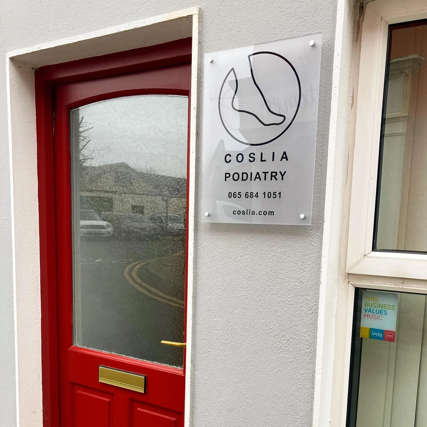 Sign for Coslia Podiatry clinic mounted on a white wall next to a red door with a glass panel and yellow mail slot. Coslia Podiatry Ennis clinic on Barrack Street in Ennis, Co. Clare.