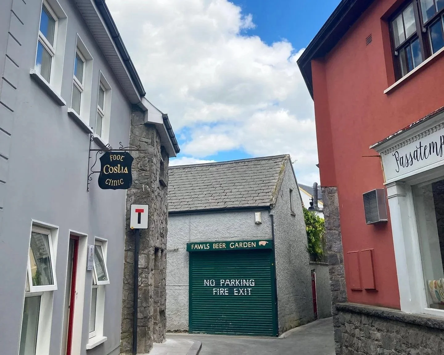 A small gray building with a sign that reads 'FAWLS BEER GARDEN' next door to Coslia Podiatry Ennis is surrounded by colorful buildings on a cloudy sky day.