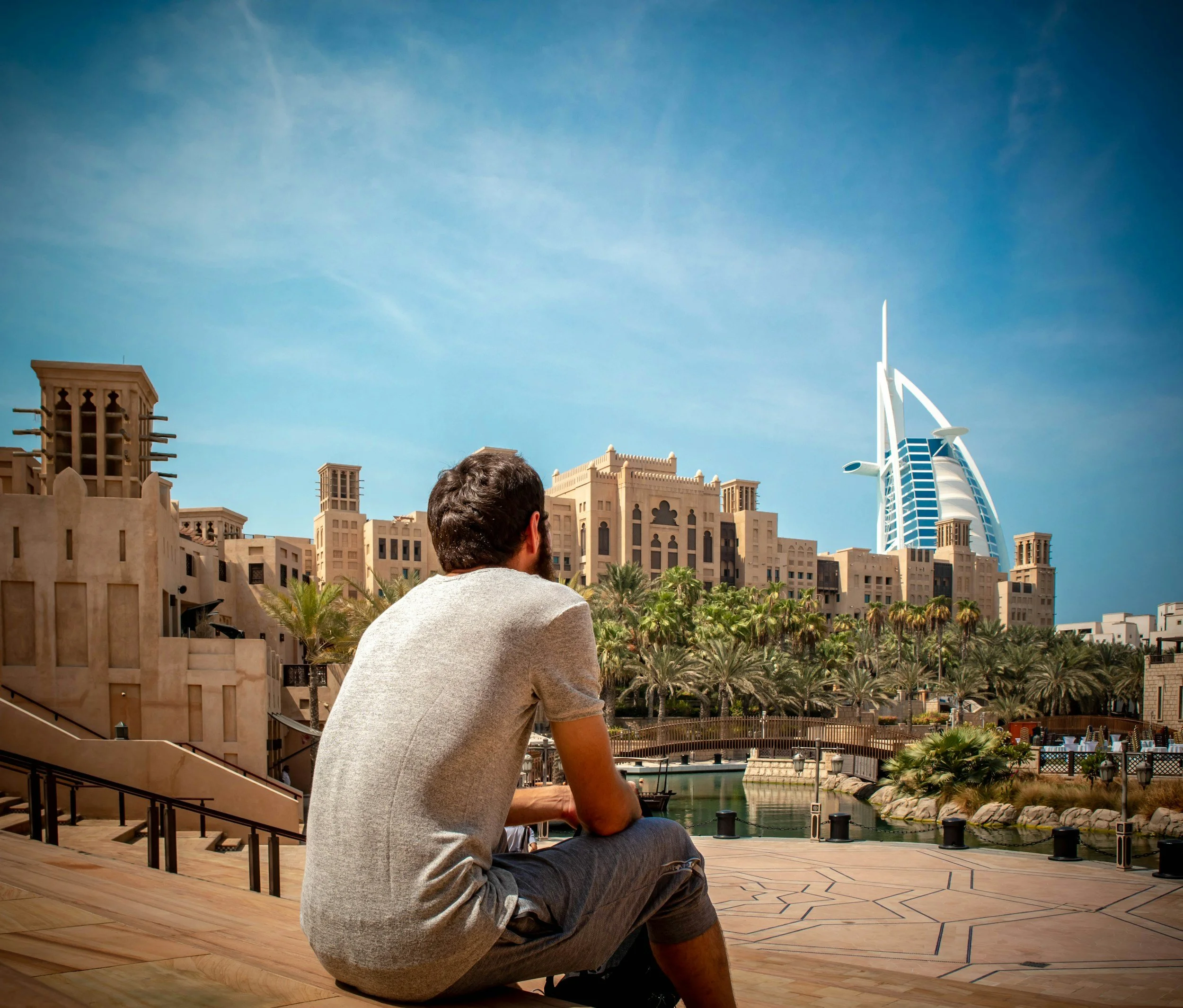 A man sitting on a wooden deck overlooking a cityscape with traditional Middle Eastern architecture, palm trees, and the Burj Al Arab hotel in Dubai under a clear blue sky.