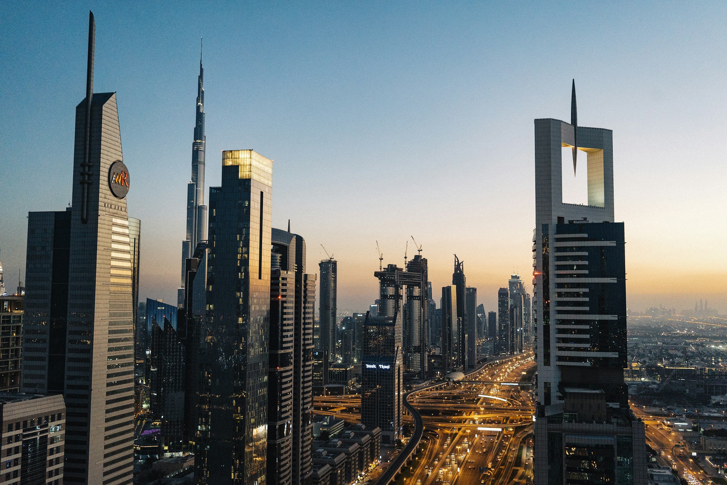 City skyline during sunset with tall skyscrapers, including the Burj Khalifa in Dubai, and busy highways with illuminated streetlights.