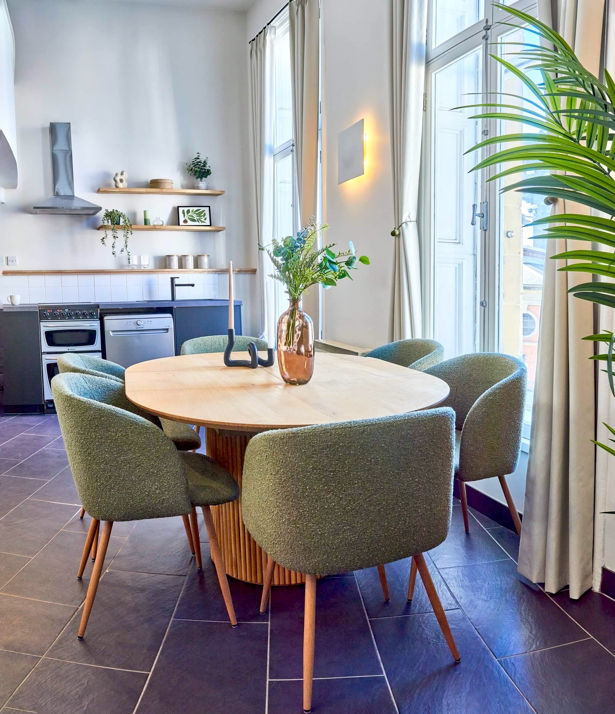 Dining room with a round wooden table, six green upholstered chairs, a vase with greenery, and natural light from large windows with white curtains.