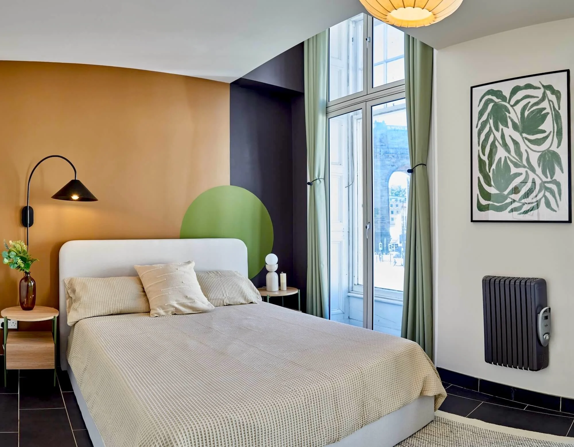 Modern bedroom with a white bed, beige bedding, and pillows. Features a black wall sconce, a small side table with a vase of flowers, large windows with green curtains, a framed leaf art, and an electric heater.