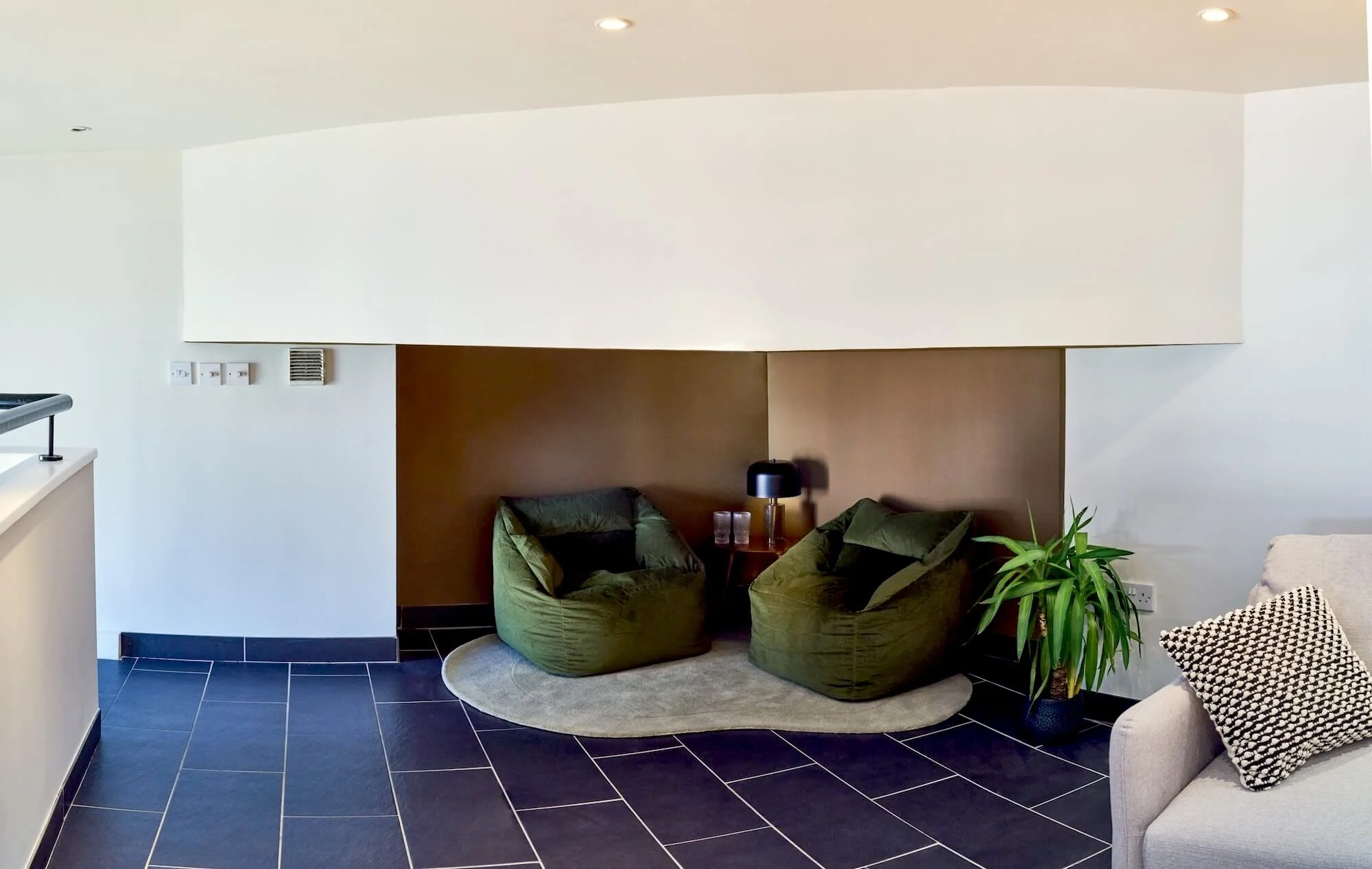 Living room with two green bean bag chairs, a small table with a black lamp, and a large potted plant, all on a gray rug against a brown wall.