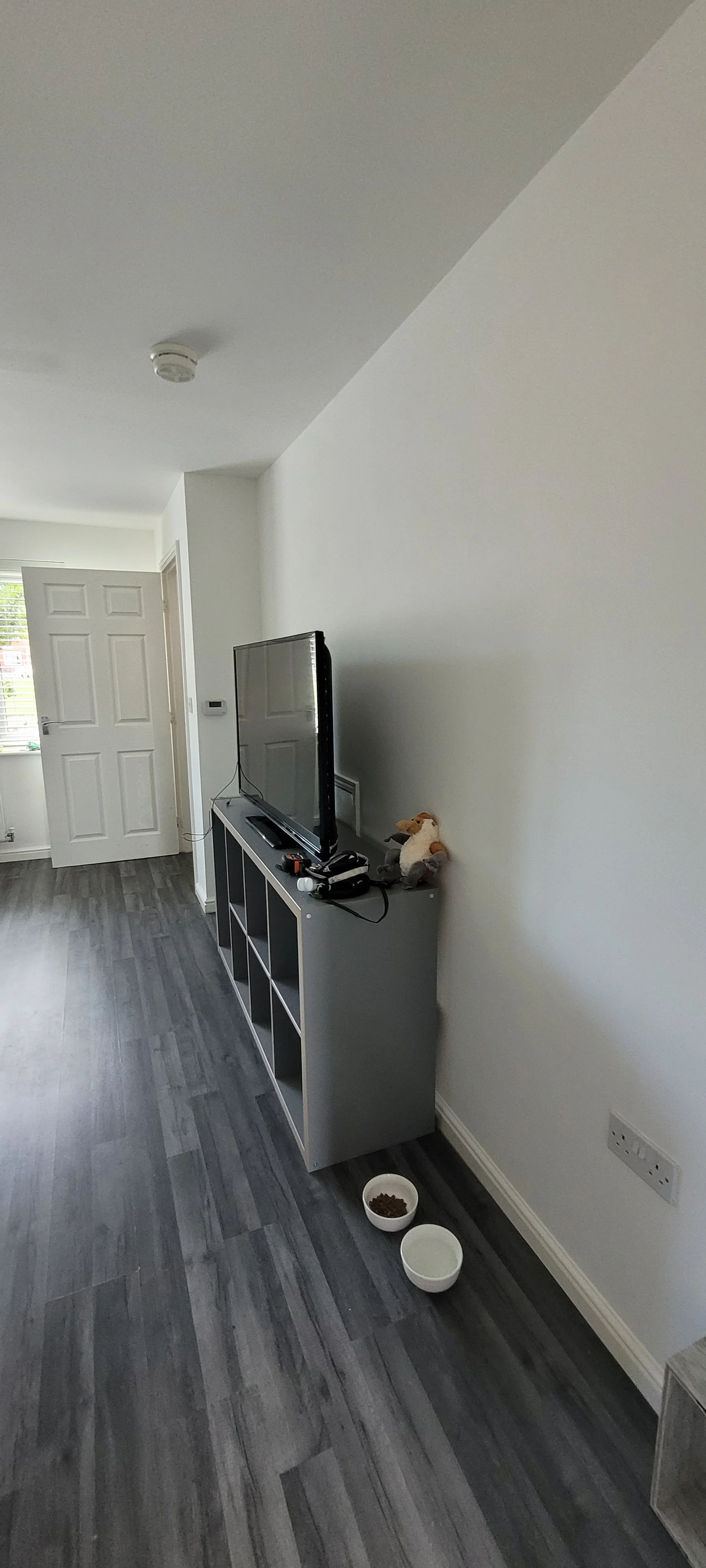 Living room with grey hardwood flooring, a white wall, and a white door. A TV is on a white TV stand with cubby holes, next to a stuffed animal and electronics. Two bowls, one with pet food, sit on the floor near the wall.