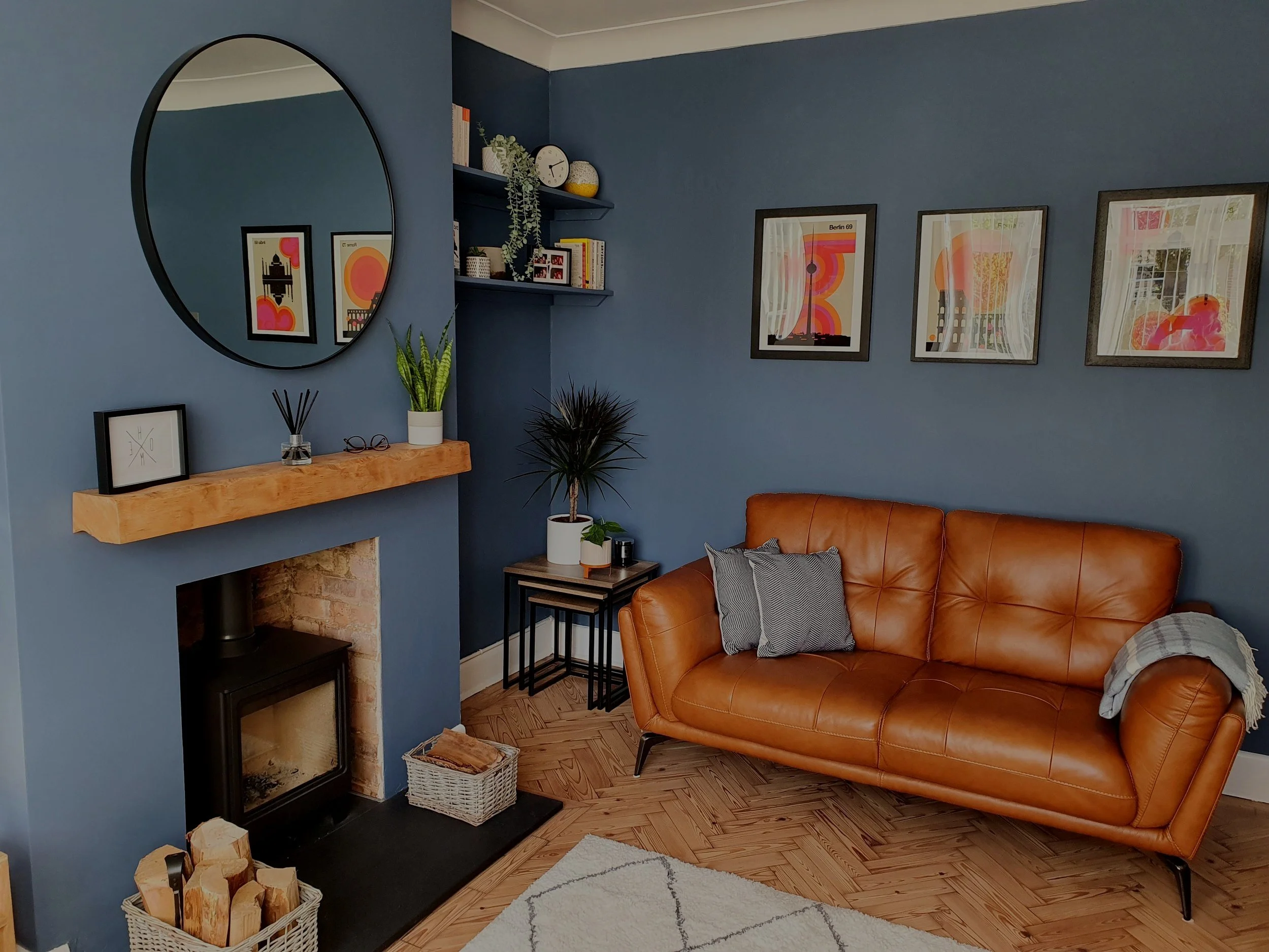 Living room with blue walls, a leather sofa, framed wall art, a fireplace with a mantel decorated with plants and glasses, a large circular mirror, and a small side table with potted plants, a black clock, and a basket of firewood.