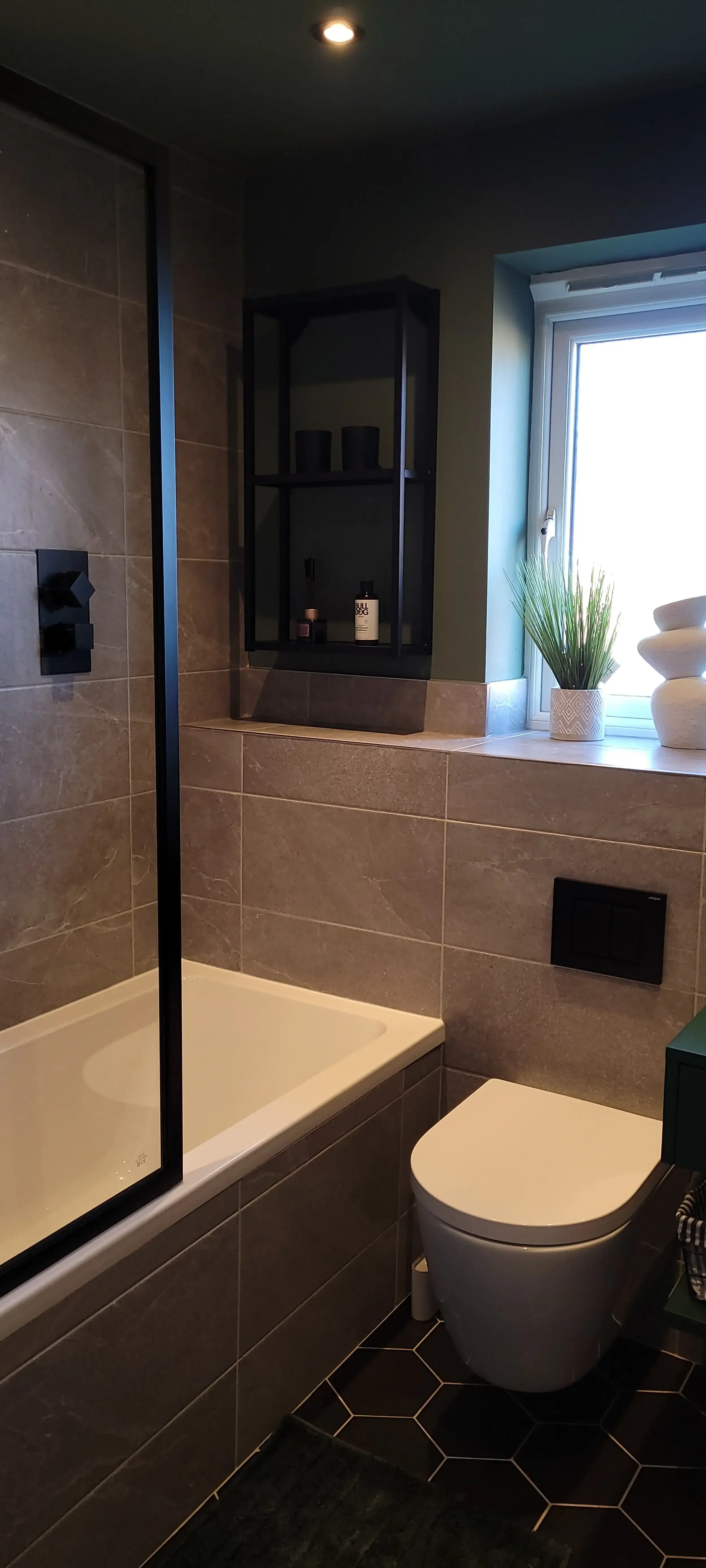 A modern bathroom featuring a bathtub with a black frame glass partition, a white toilet, a window with a potted plant and decorative stones, and dark hexagonal floor tiles.