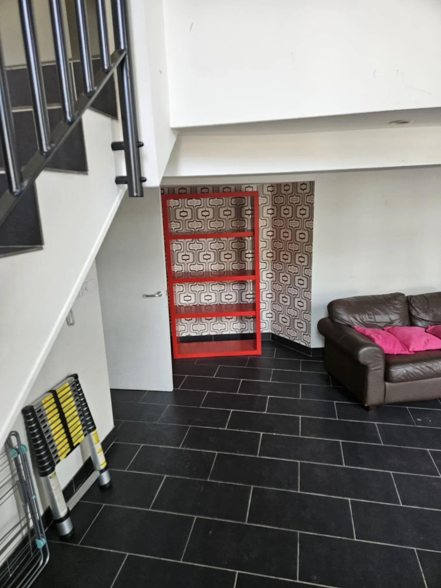 A living room with dark tiled flooring, a brown leather sofa with pink pillows, a patterned wall, a red metal shelving unit, a closed white door, and part of a staircase with black railing.