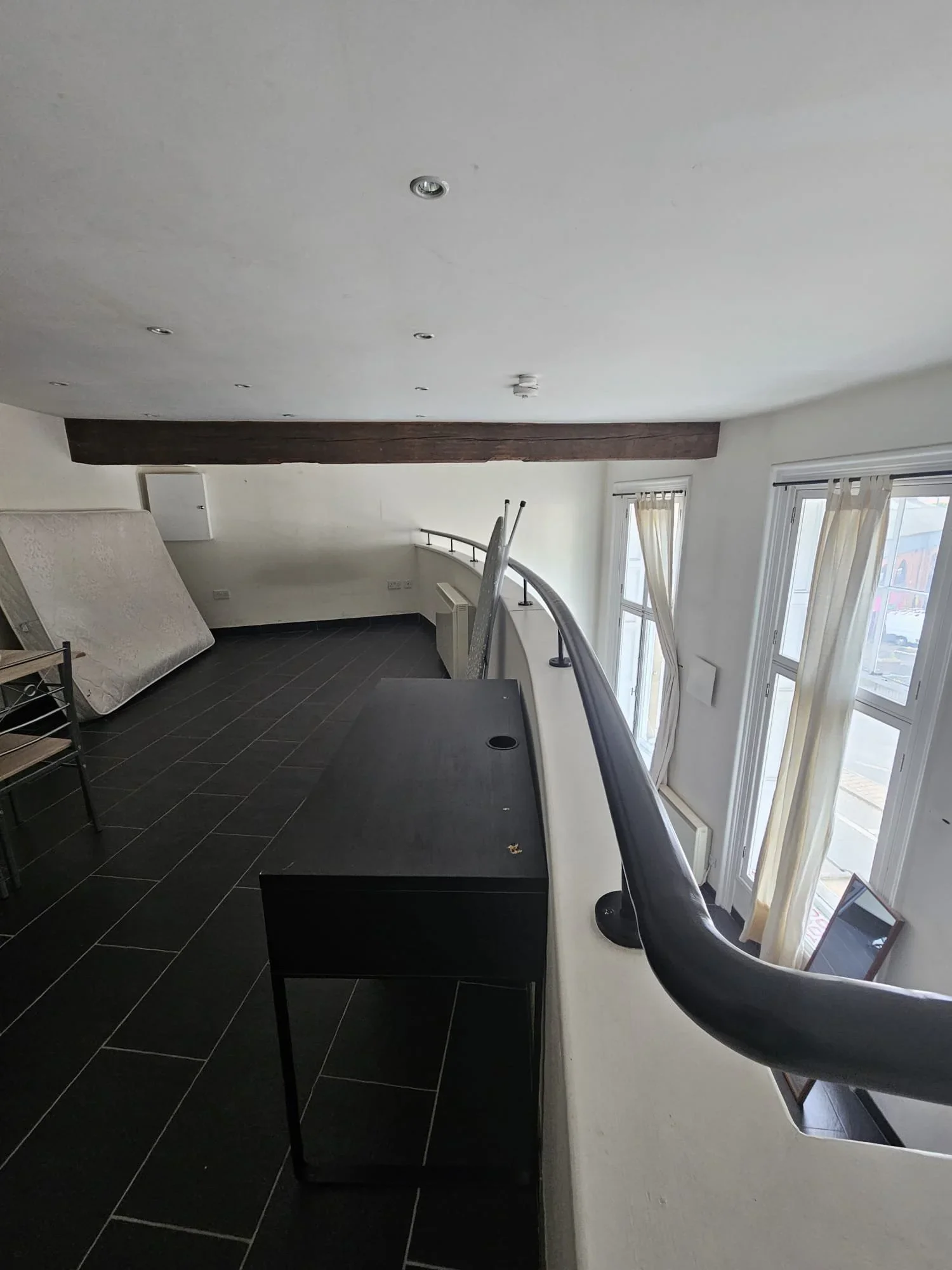 Interior view of an empty room with black tiled flooring, large windows with beige curtains, a curved white wall with a black handrail, and some furniture including a small black table and a mattress leaning against the wall.