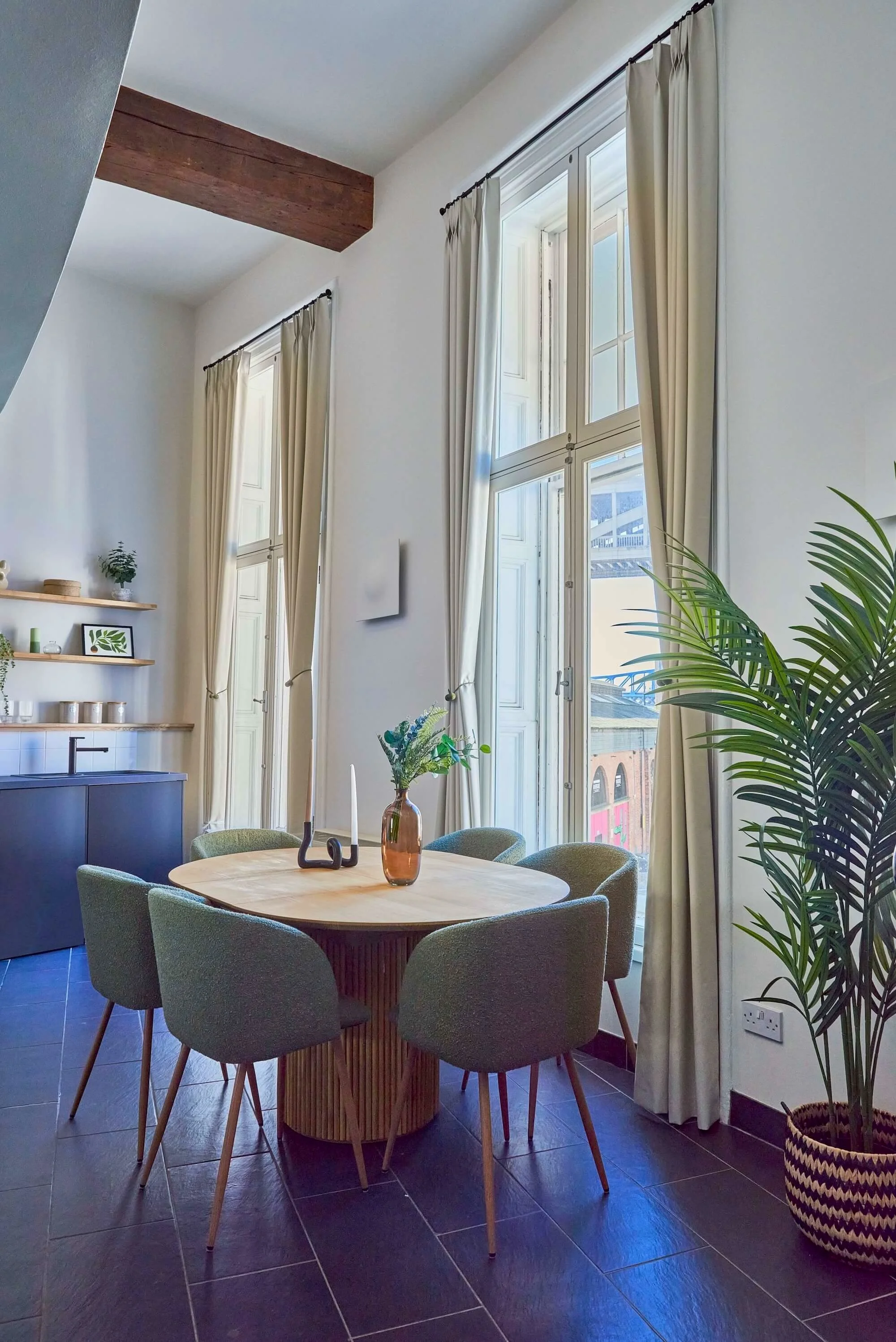 A dining area with a round wooden table surrounded by six green upholstered chairs, a large window with white framing and cream curtains, a potted plant in a woven basket, and shelves with decorative items in the background.