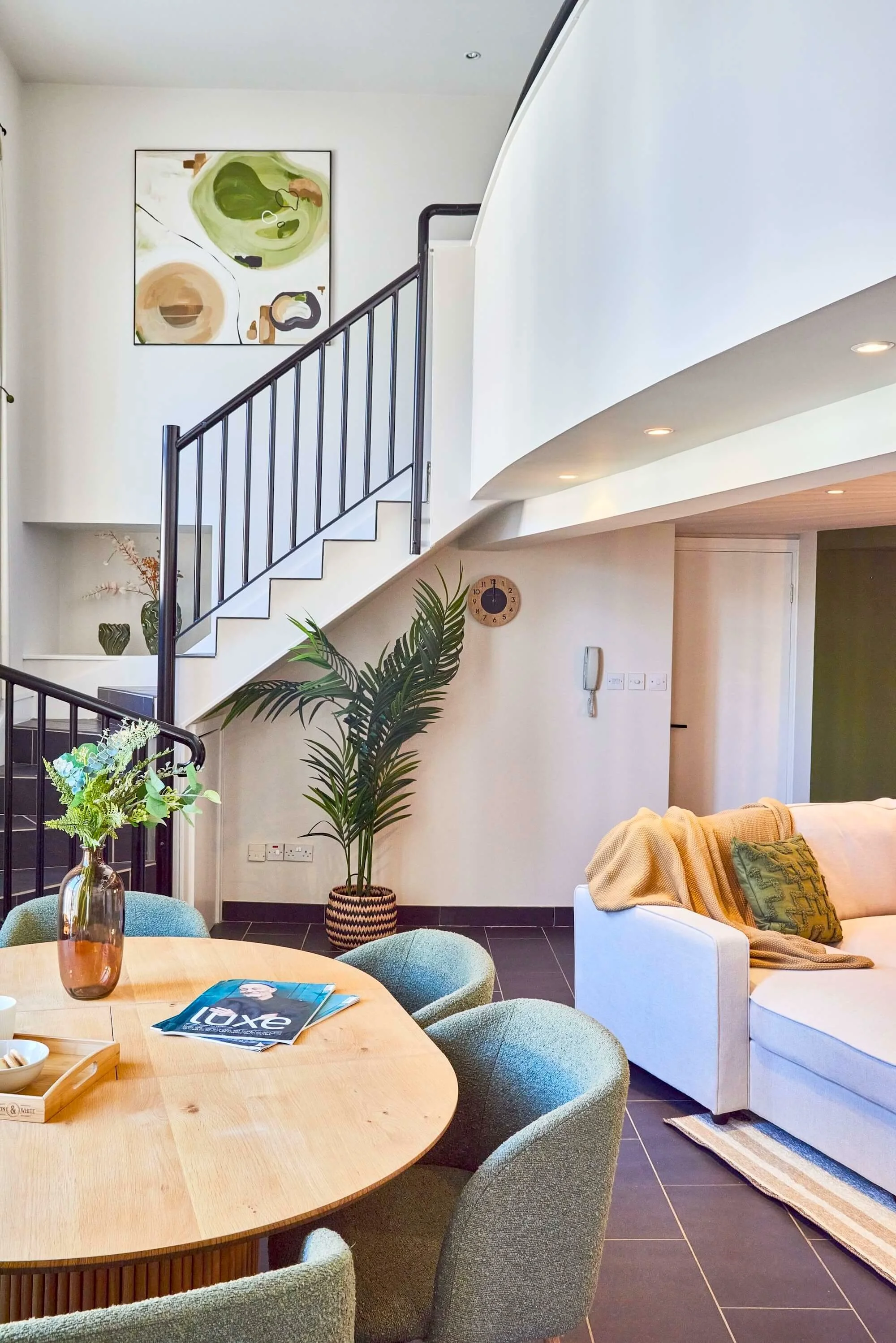 A modern living room with a staircase, sitting area, and decorative elements including a large plant and artwork on the walls.