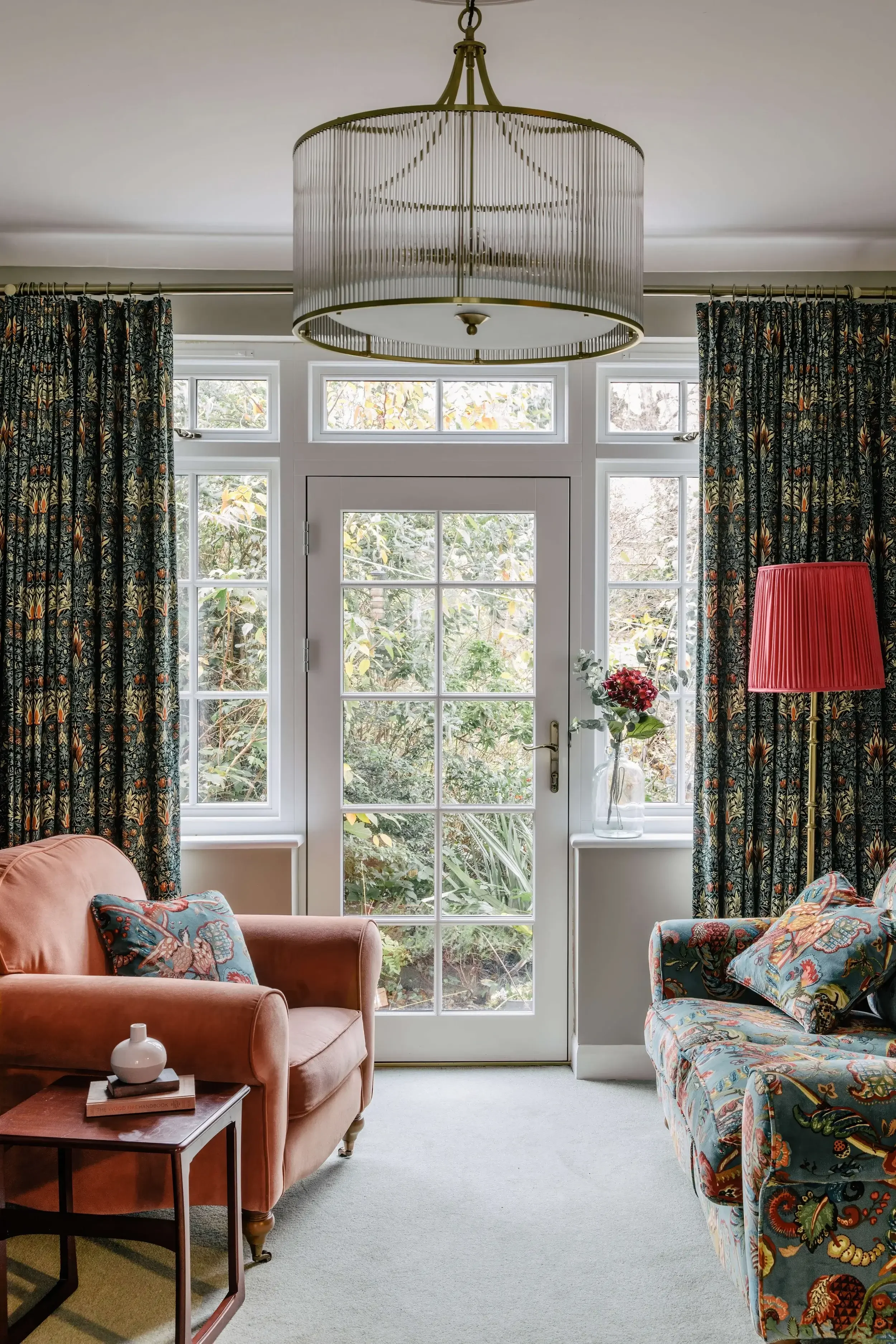 Living room with a glass-paneled door leading to a garden, floral curtains, a pinkish-orange armchair, a patterned sofa, a small wooden side table with a white vase, and a red lampshade.