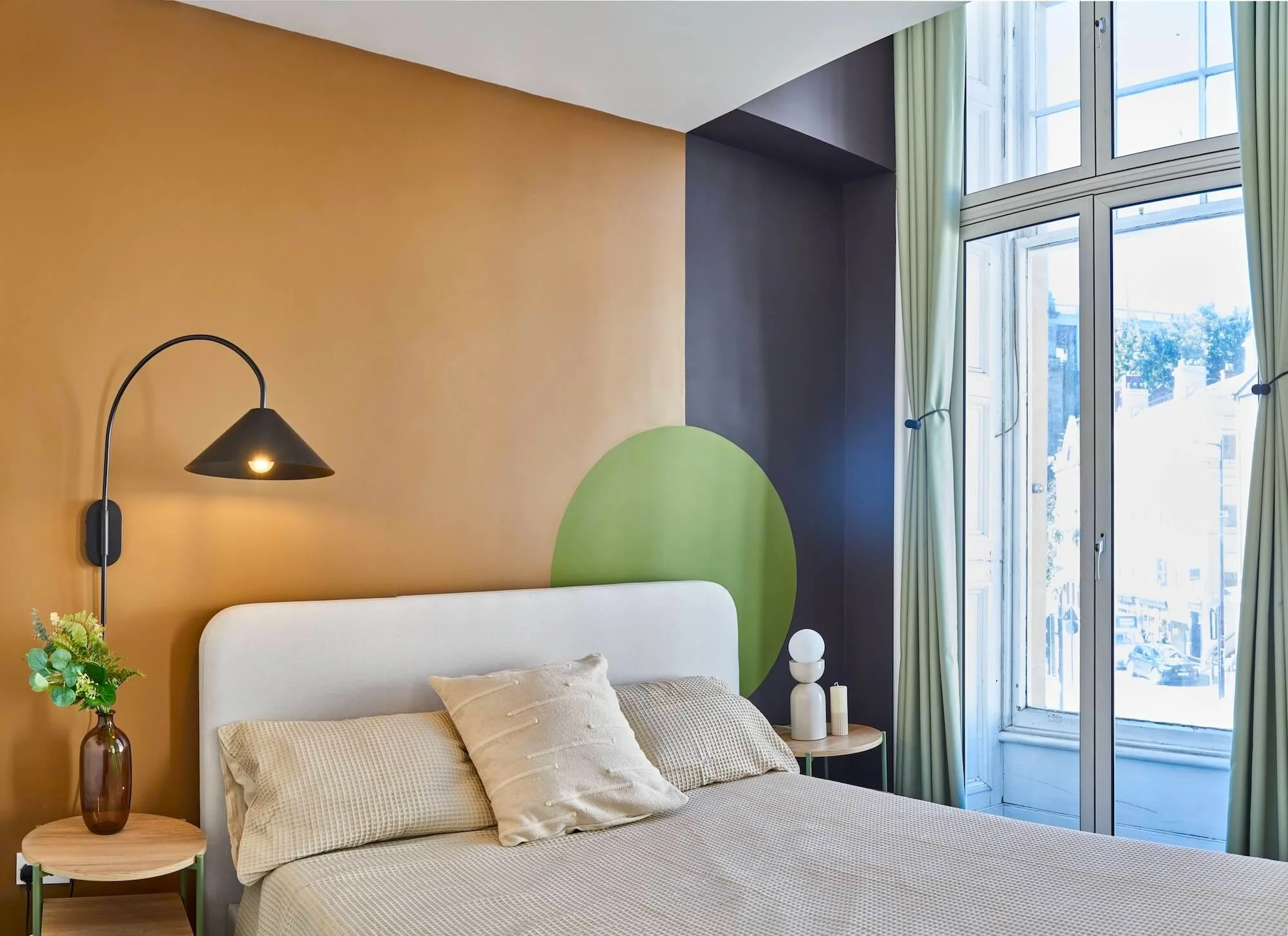 Bedroom with beige bedding, white headboard, green accent wall with circle design, black wall lamp, small wooden side table with vase and flowers, large window with light green curtains, and a modern nightstand with white decorations.
