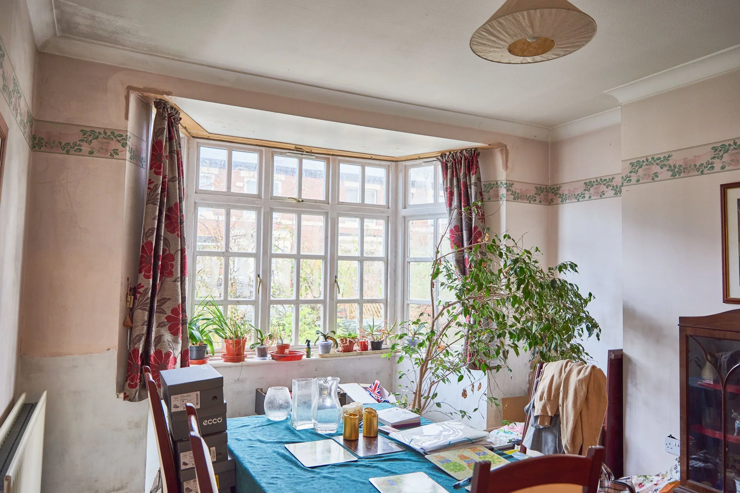 A dining room with a large bay window, red and beige curtain, potted plants on the windowsill, a table with various items including glasses, books, and candles, and a large leafy plant next to the table.