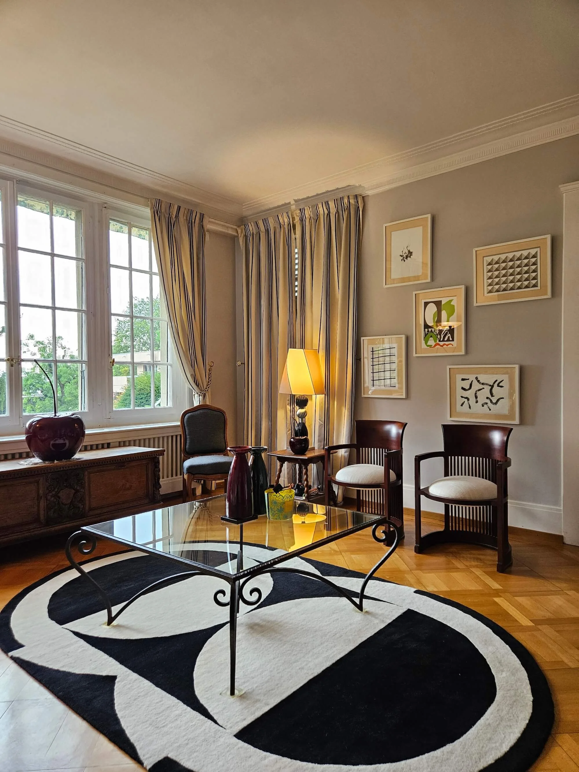 Living room with two windows, striped curtains, a table with vases, and several framed artworks on the wall.