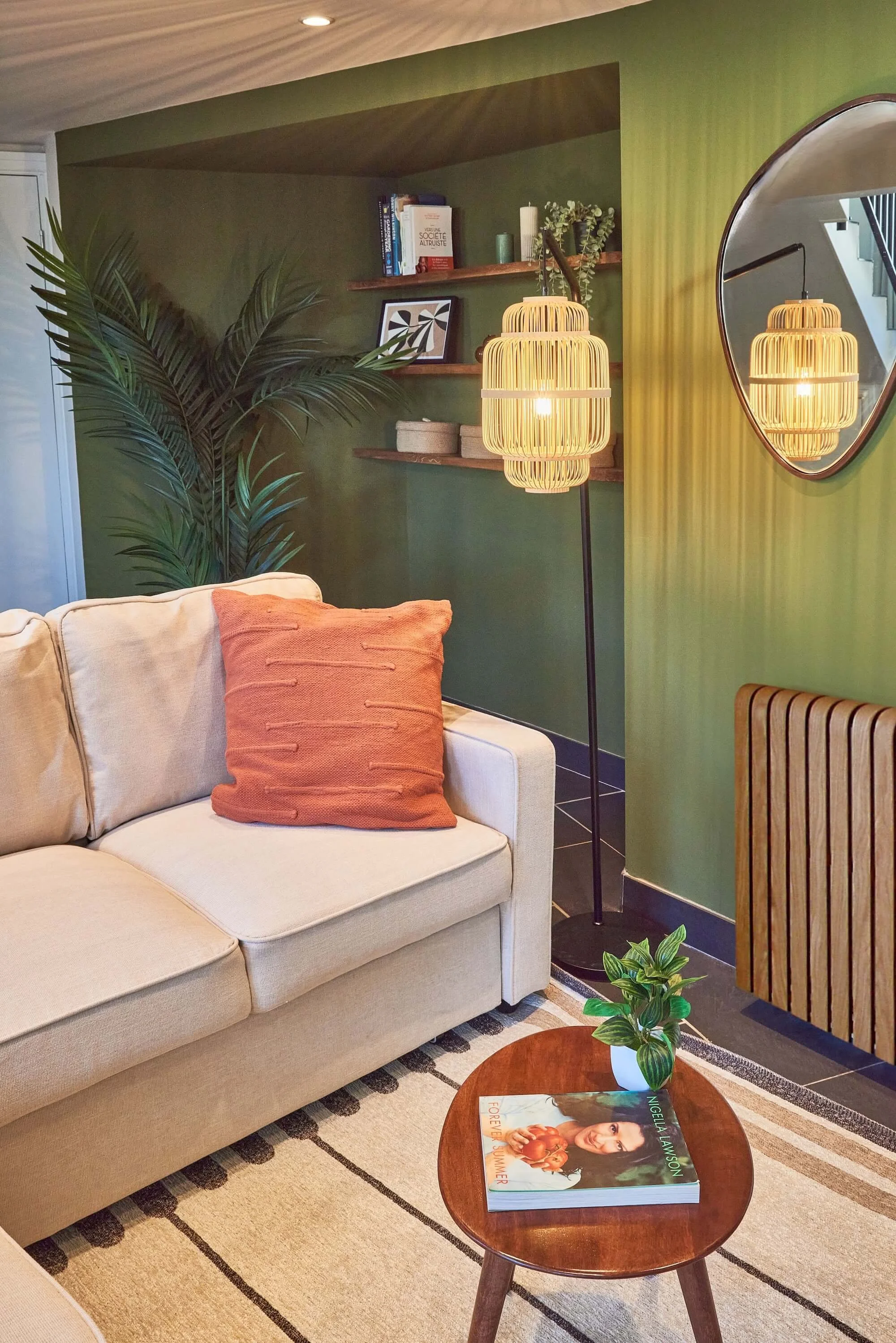 Living room with a beige sofa, orange cushion, wooden side table with a book and potted plant, green wall with shelves, mirror, and a tall floor lamp with a woven shade, large potted plant in the corner.
