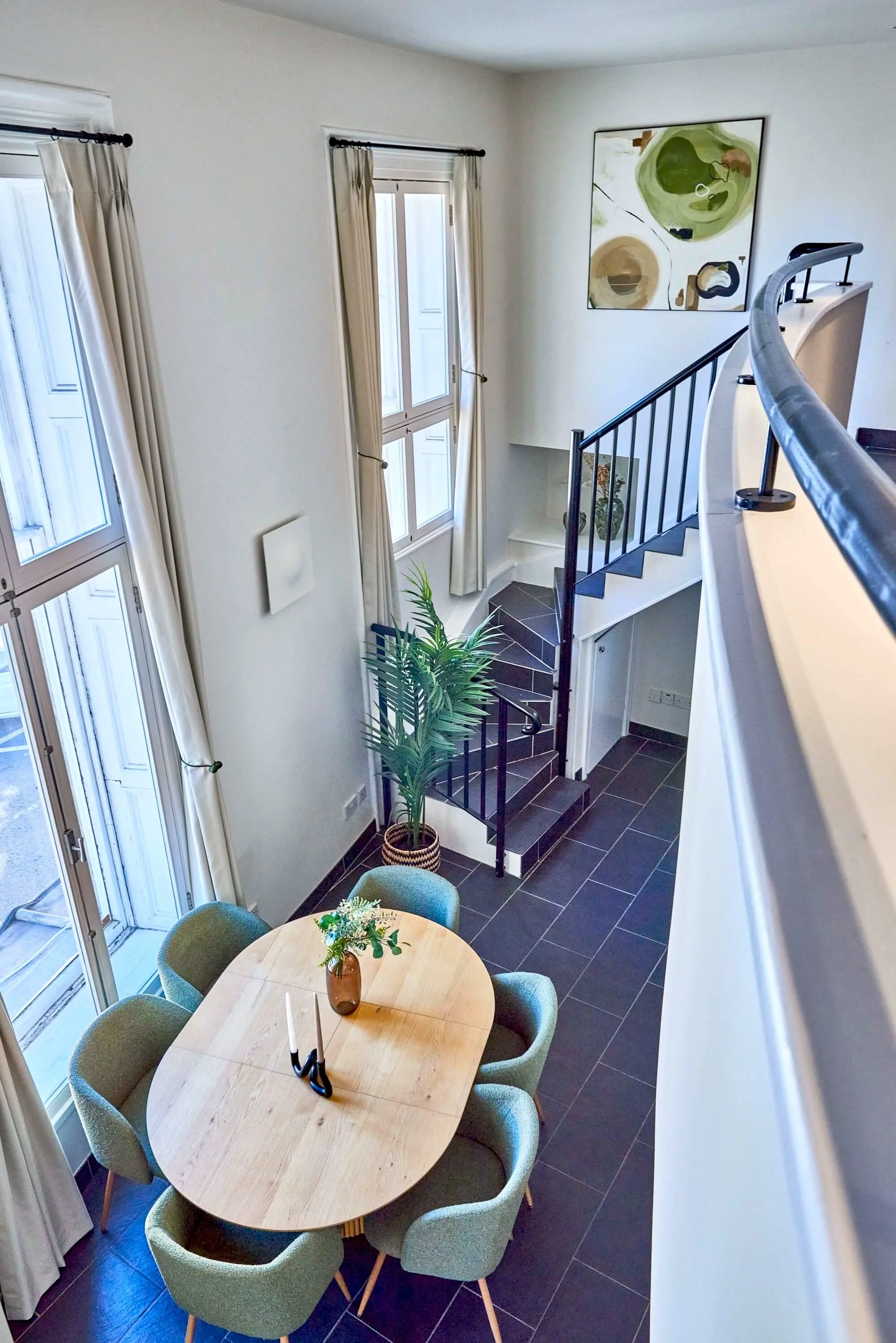 View of a living and dining area with tall windows, beige curtains, a round wooden dining table with six green chairs, potted plants, a staircase with black railing, and a large abstract painting on the wall.