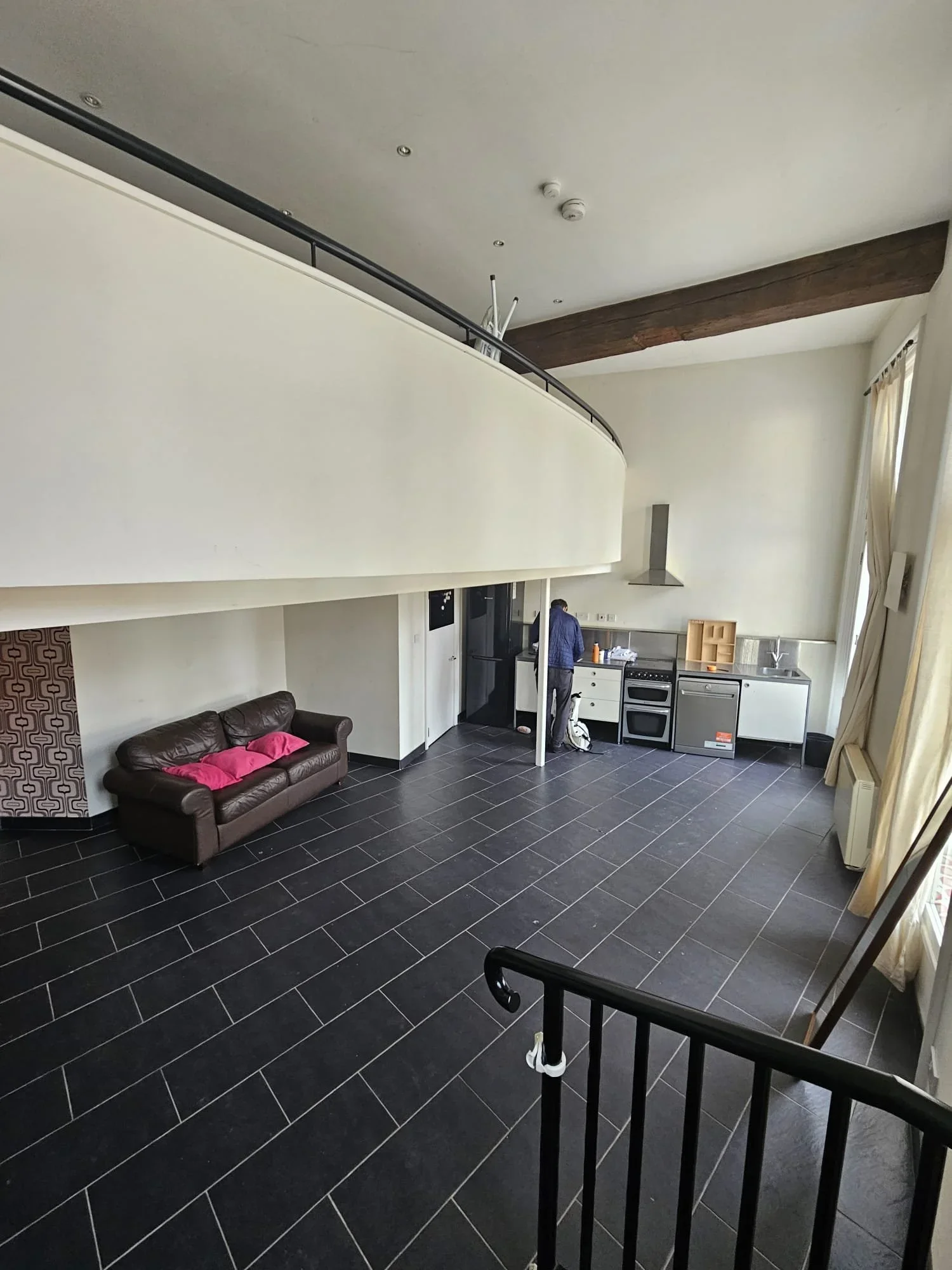 Spacious room with black tiled floor, a small kitchen area, a brown leather couch with pink cushions, and a staircase railing in the foreground.