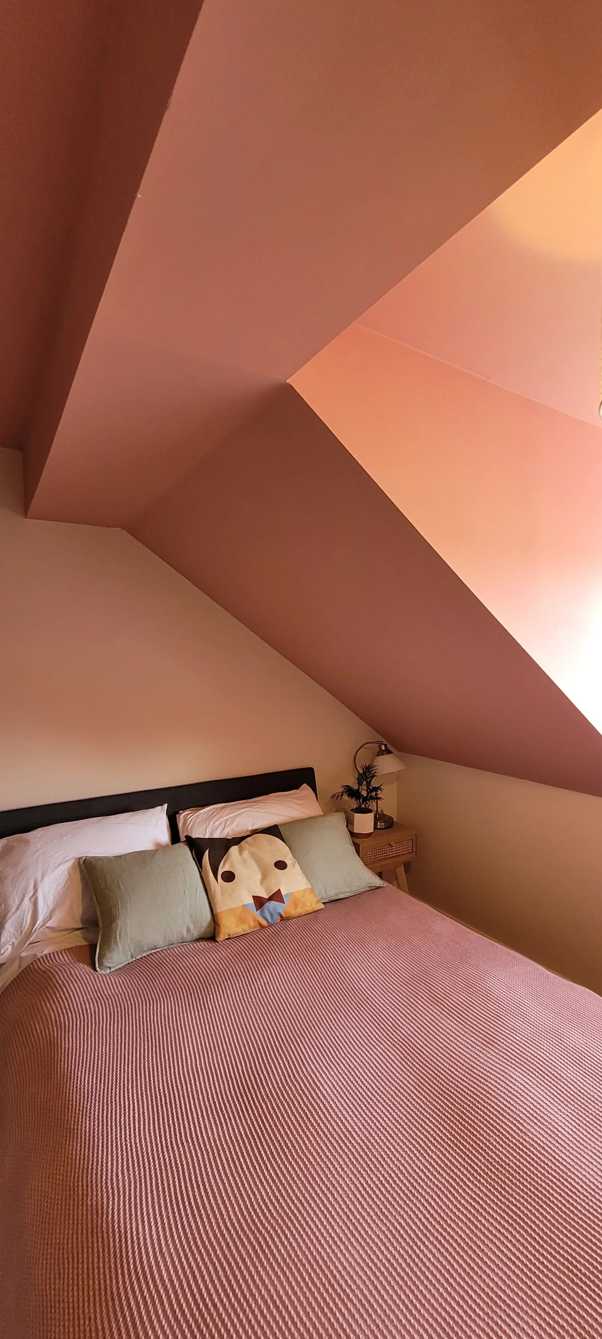 A bedroom with a sloped ceiling and a bed with white sheets, green pillows, and a decorative pillow with a dog's face. A small bedside table with a lamp and plant are beside the bed.