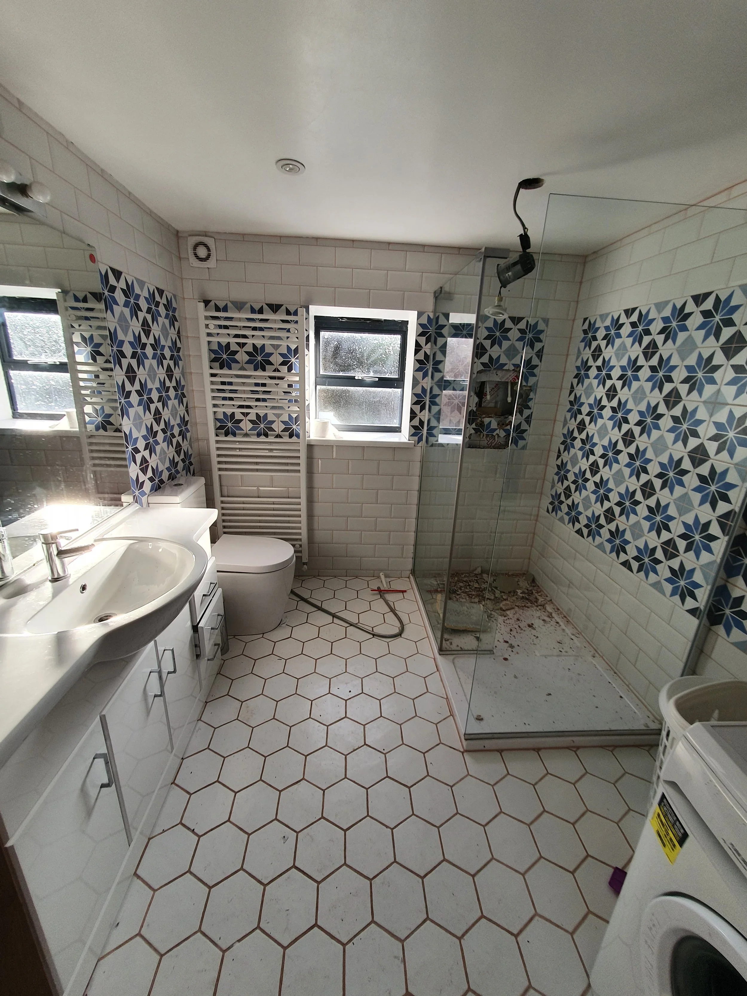 A bathroom with white hexagonal floor tiles accented with brown grout, a white sink with a mirror above, a modern toilet, a heated towel rail, a window, and a glass-enclosed shower with some dirt or debris inside.