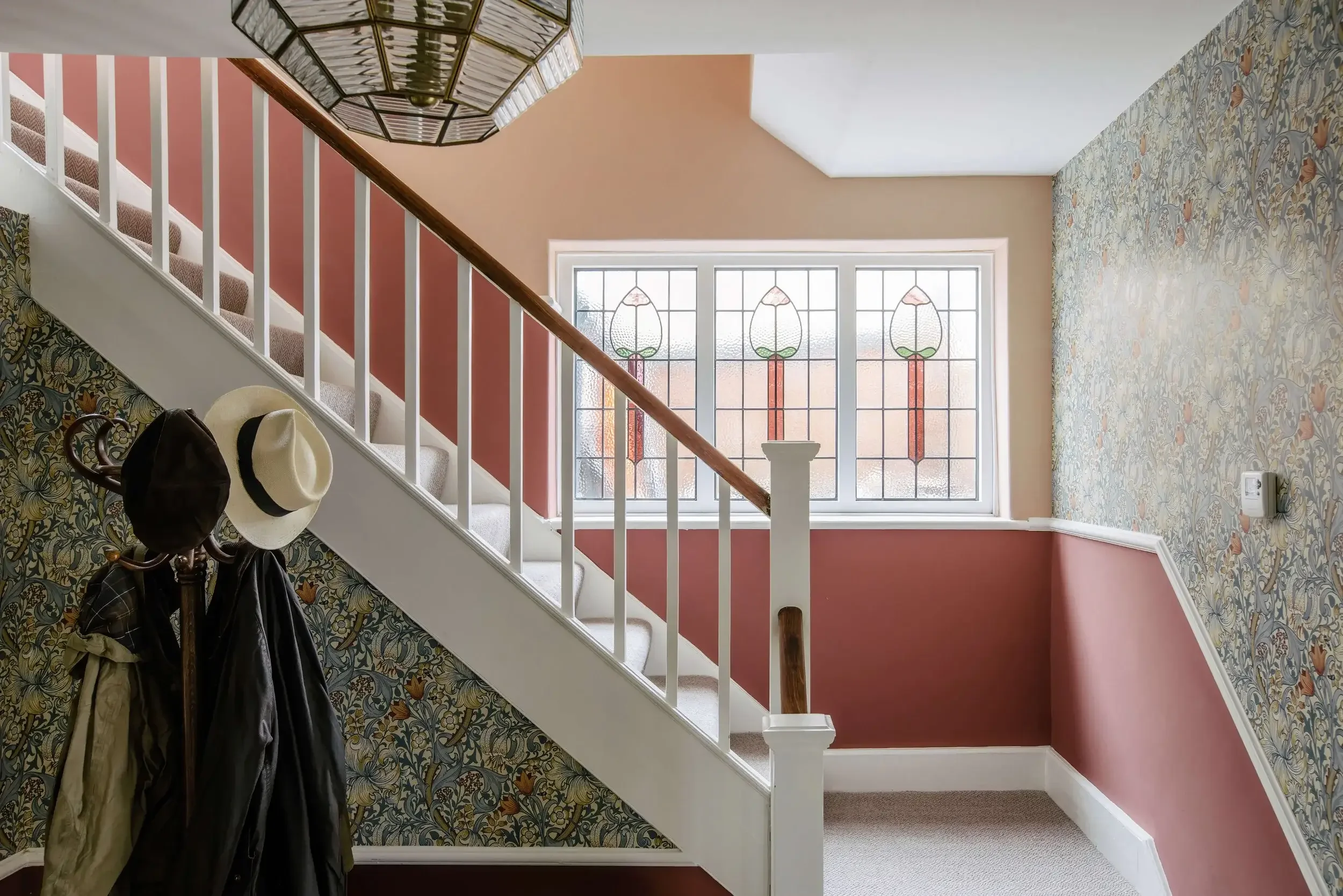 Interior of a house with a staircase, patterned wallpaper, stained glass window, and coat hooks with hats and jackets.