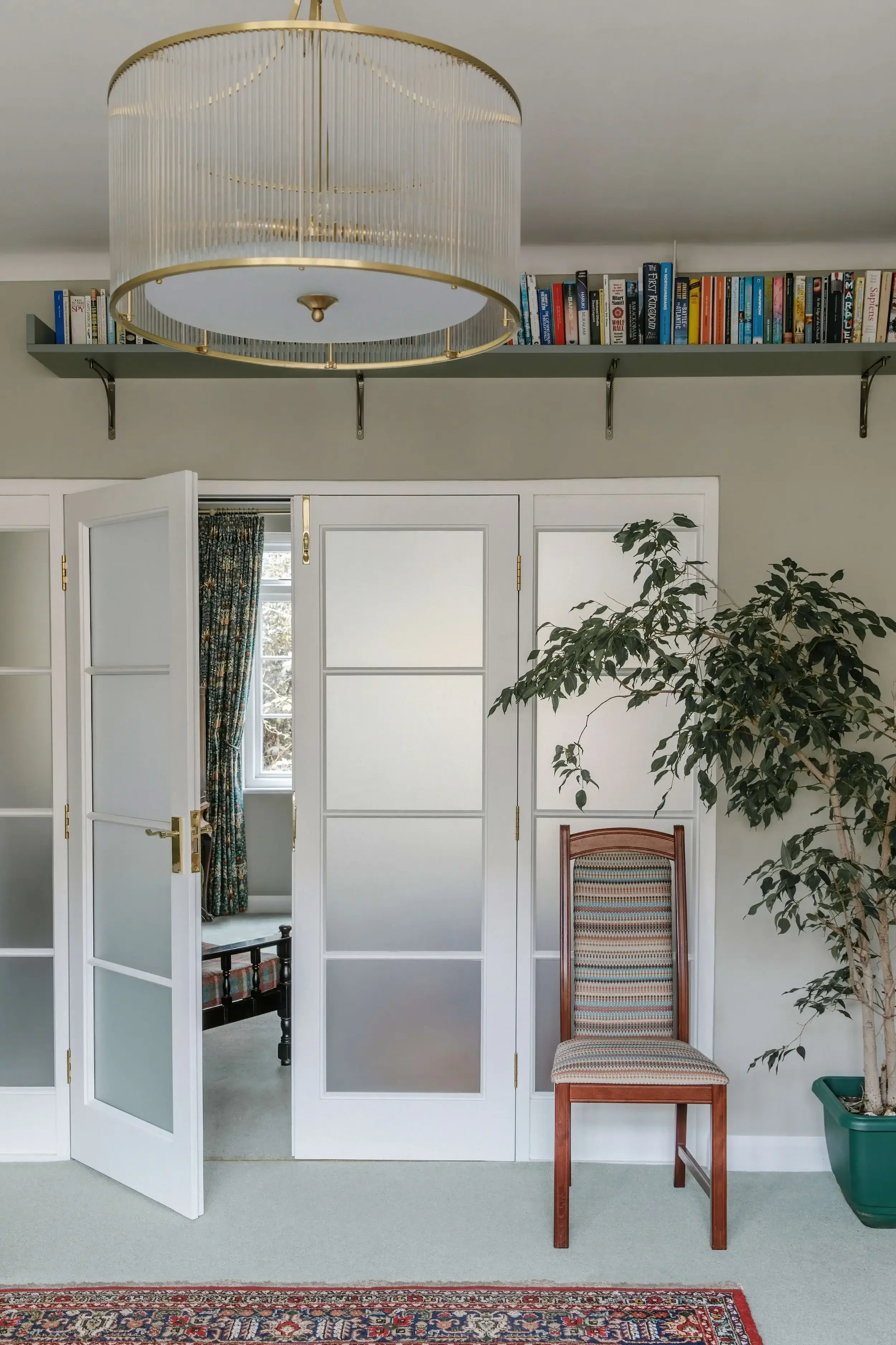 Interior view of a room with a large circular chandelier, a bookshelf, a partially open glass door revealing a bed and window with floral curtains, a wooden chair with upholstery, a potted plant, and a colorful rug.