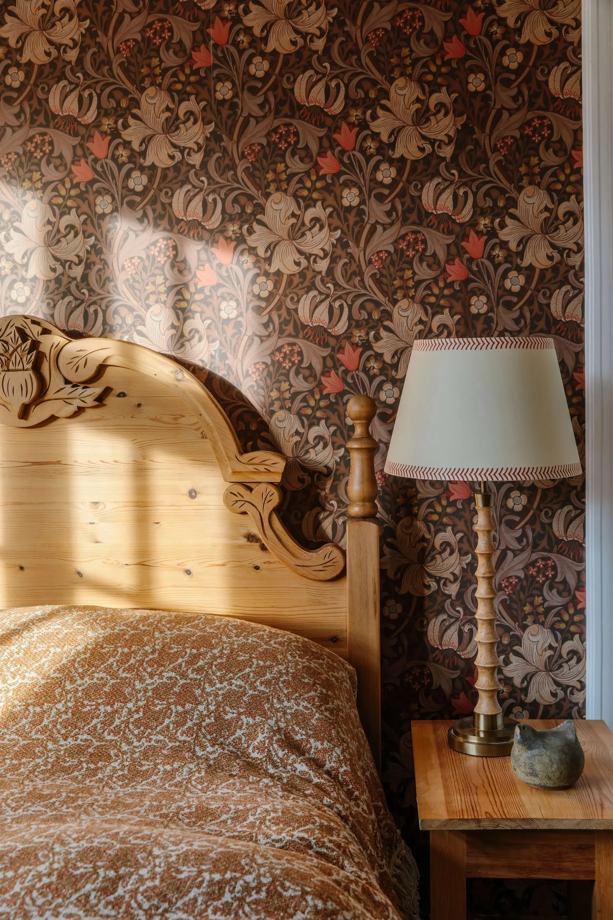 A bedroom corner with a wooden carved headboard, a patterned floral wallpaper, a beige and brown patterned bedspread, a wooden nightstand, a lamp with a decorative wooden base and a white shade, and a small stone or ceramic decorative object on the n