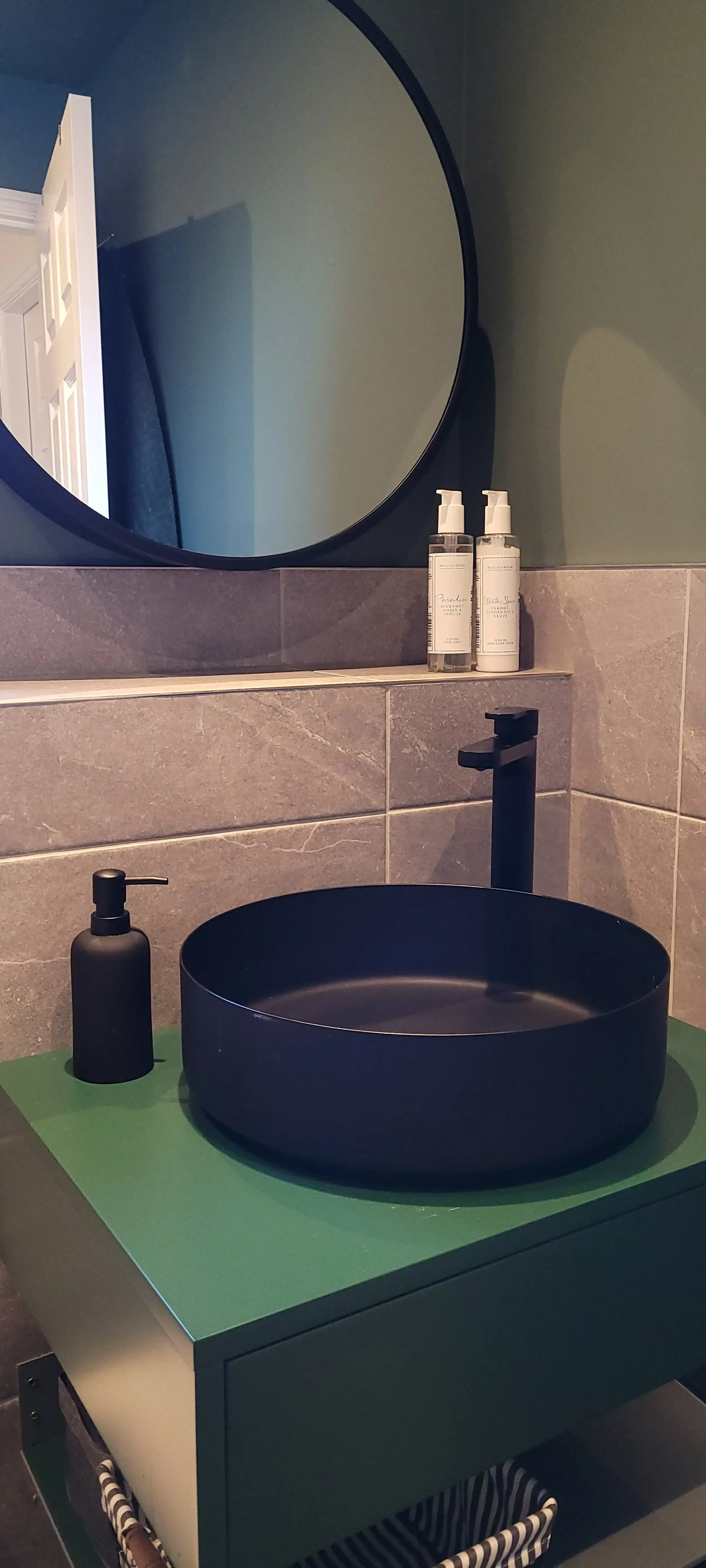 A bathroom sink with a black round vessel basin, black soap dispenser, and two bottles of skincare products on a gray tiled ledge in front of a mirror. The sink rests on a green cabinet.