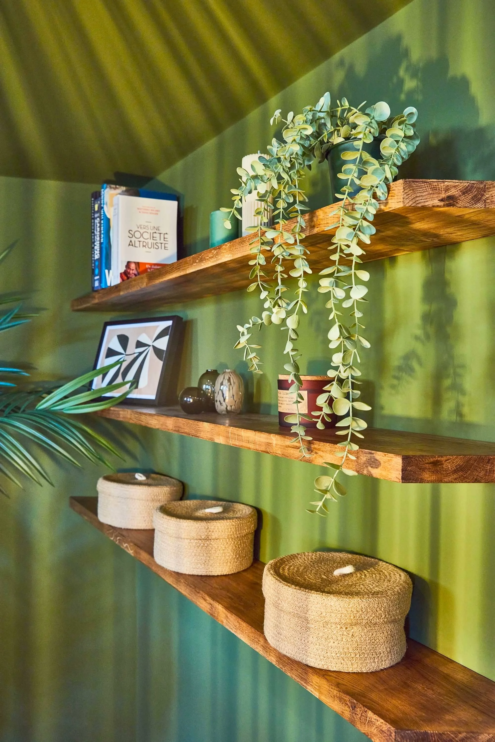 Three wooden shelves with decorative objects and plants against a green wall. The top shelf has books and a potted plant with trailing vines. The middle shelf has framed artwork and small vases. The bottom shelf has three woven baskets with lids.