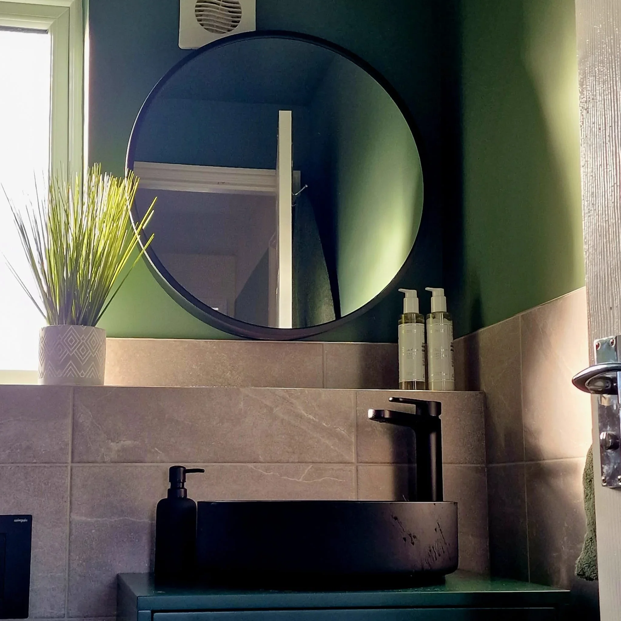 A bathroom vanity with a black vessel sink, black faucet, soap dispenser, wall-mounted mirror, green walls, a perennial plant in a white patterned pot, two pump bottles, and a wooden door.