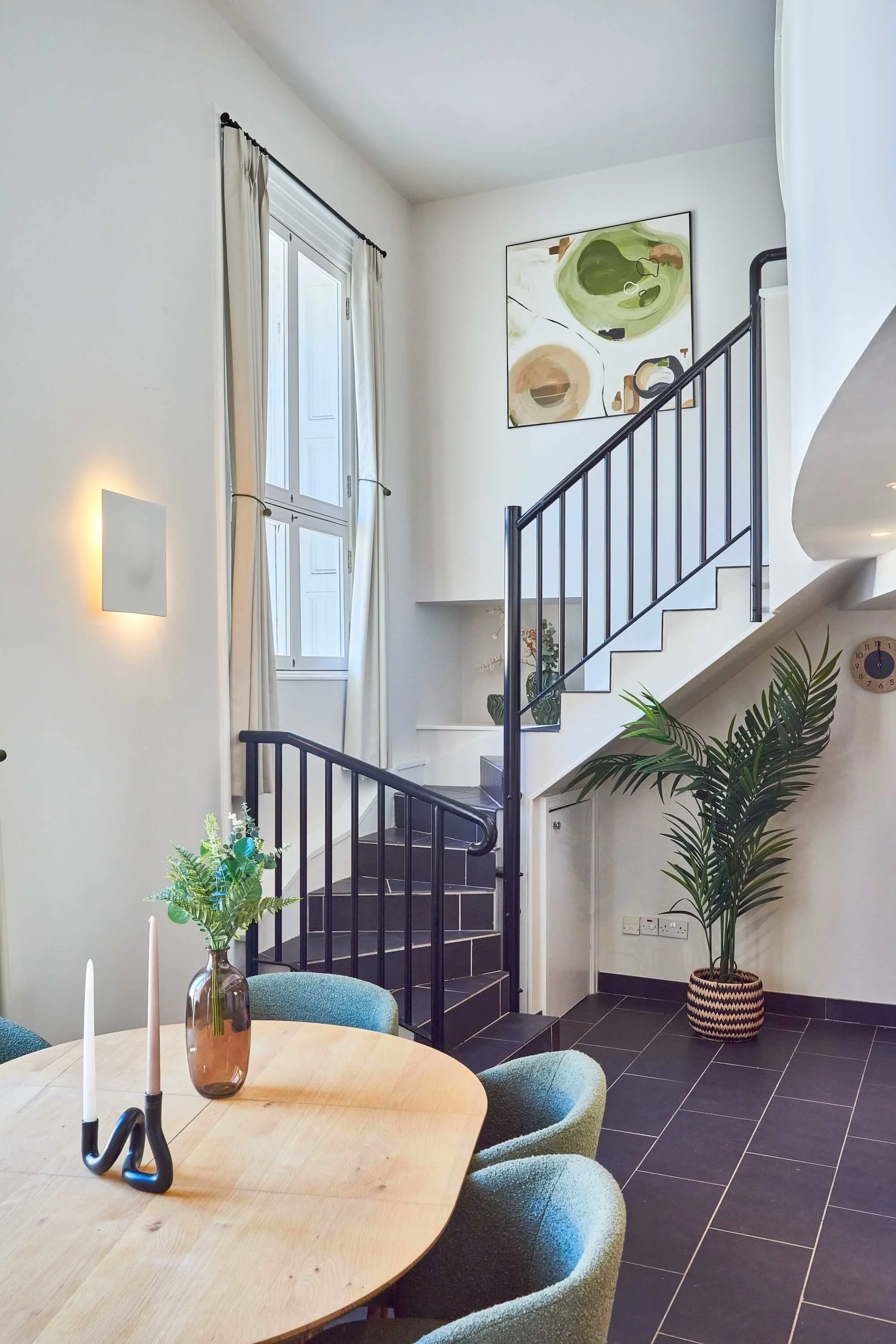 Modern living space with staircase, large window with curtains, abstract artwork on the wall, potted plants, dining table with candles and a vase, black tile flooring, and minimalist decor.