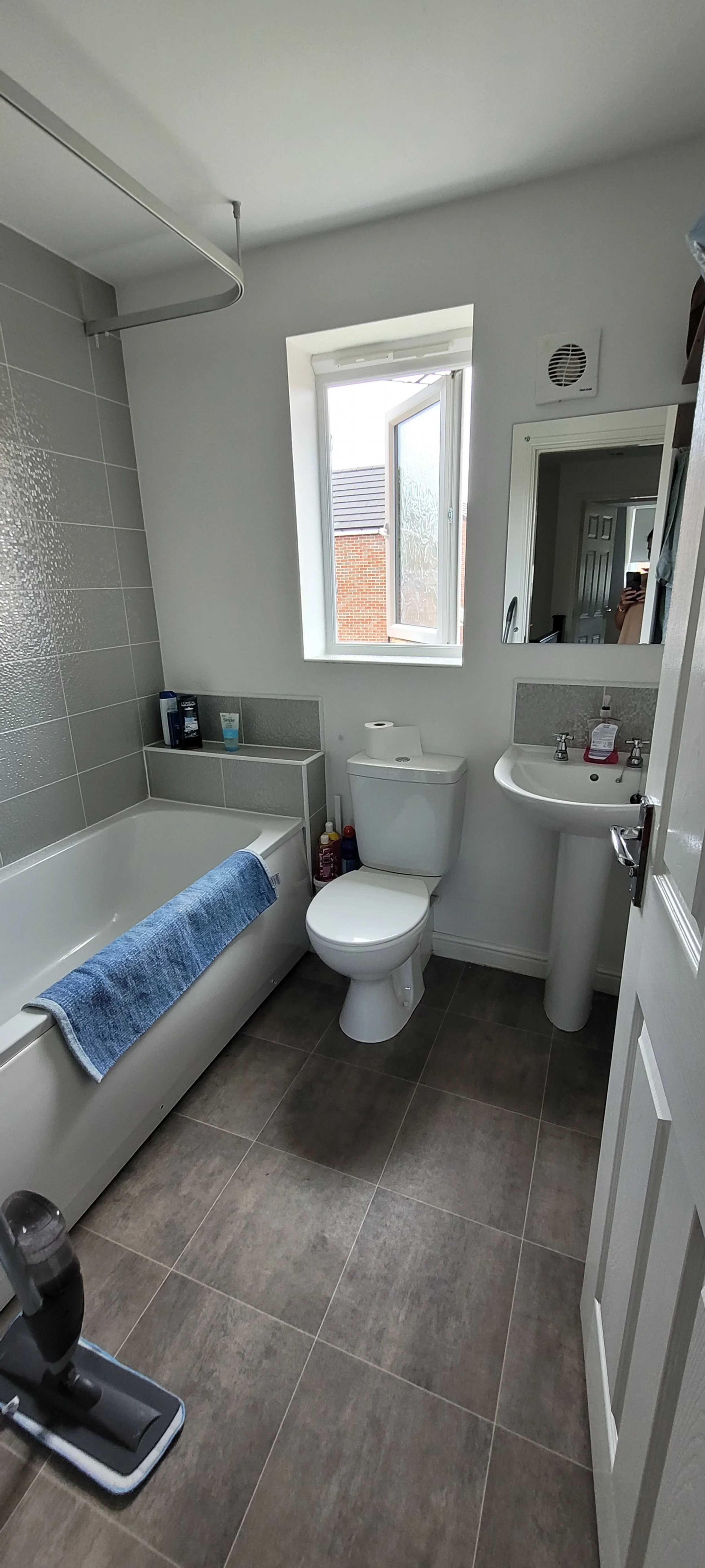 A small bathroom with a bathtub, toilet, pedestal sink, and a window open to the outside. The bathtub has a blue towel hanging over the edge, and various toiletries are placed on the ledge and on the floor.