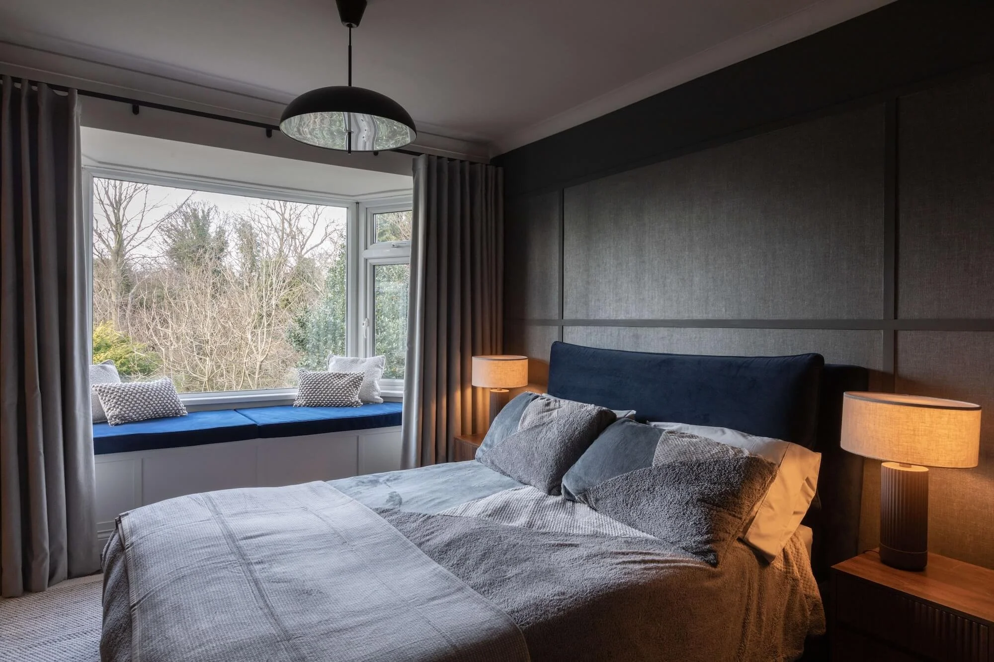 A bedroom with a large bed, dark blue headboard, two nightstands with lamps, a window seat with pillows, and a view of trees outside.