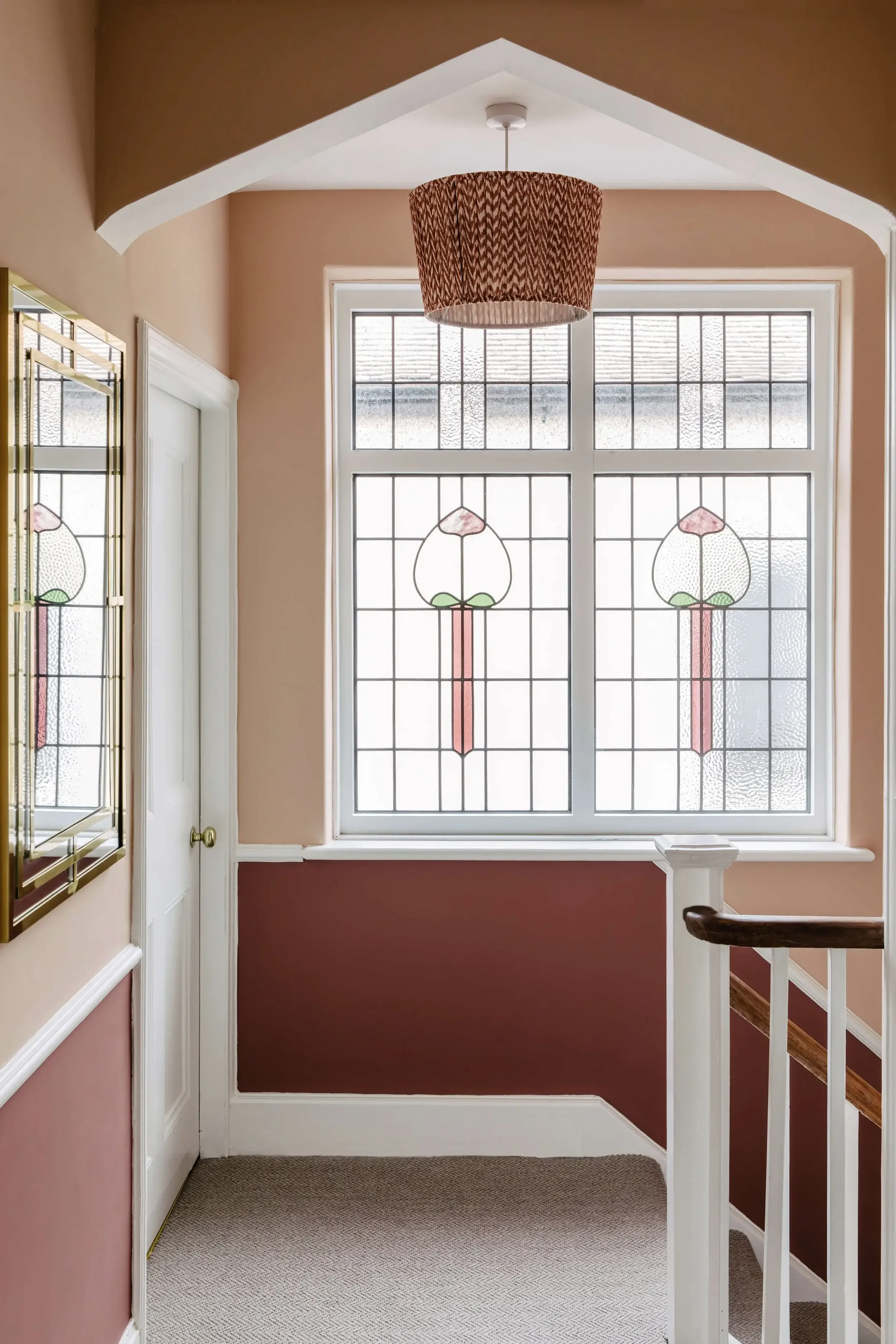 A staircase landing with a large stained glass window featuring pink flowers and green leaves, a ceiling light with a patterned shade, and pastel-colored walls.