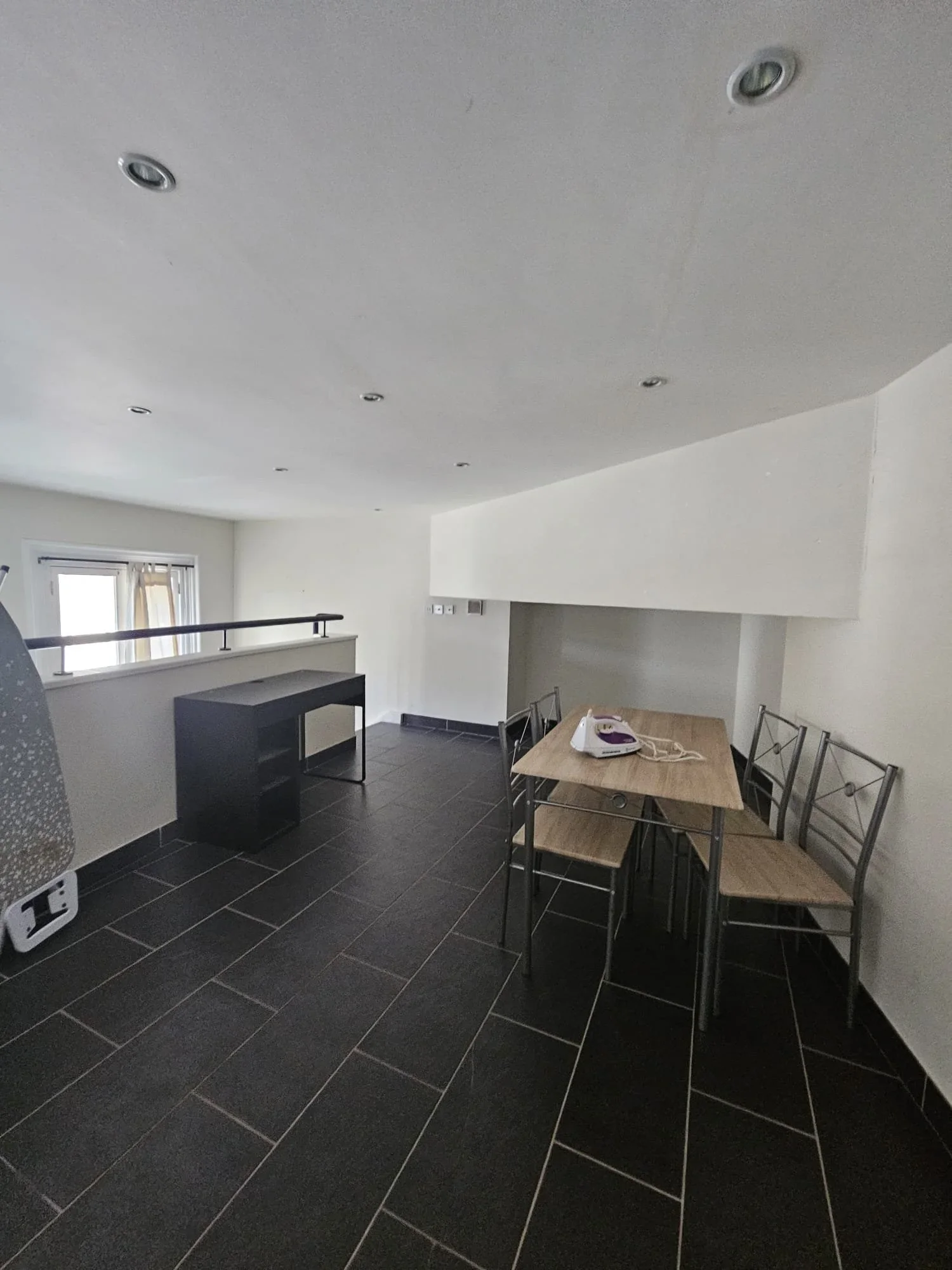 Empty room with dark tiled floor, white walls, a small dining table with four chairs, and a black desk near a window.