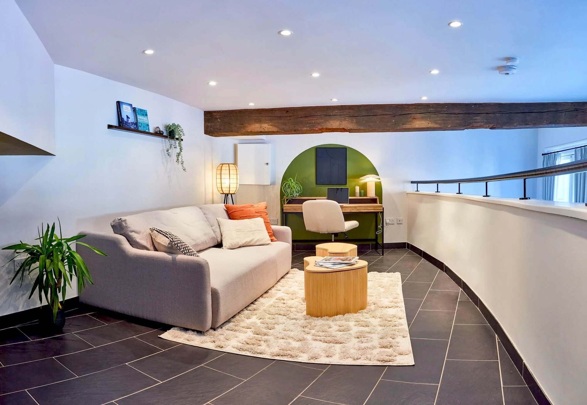 Modern living room with a beige sofa, wooden coffee tables, a potted plant, and a small desk with a white chair. The room features white walls, a green accent wall, and a large wooden ceiling beam, with recessed lighting and a fluffy area rug.