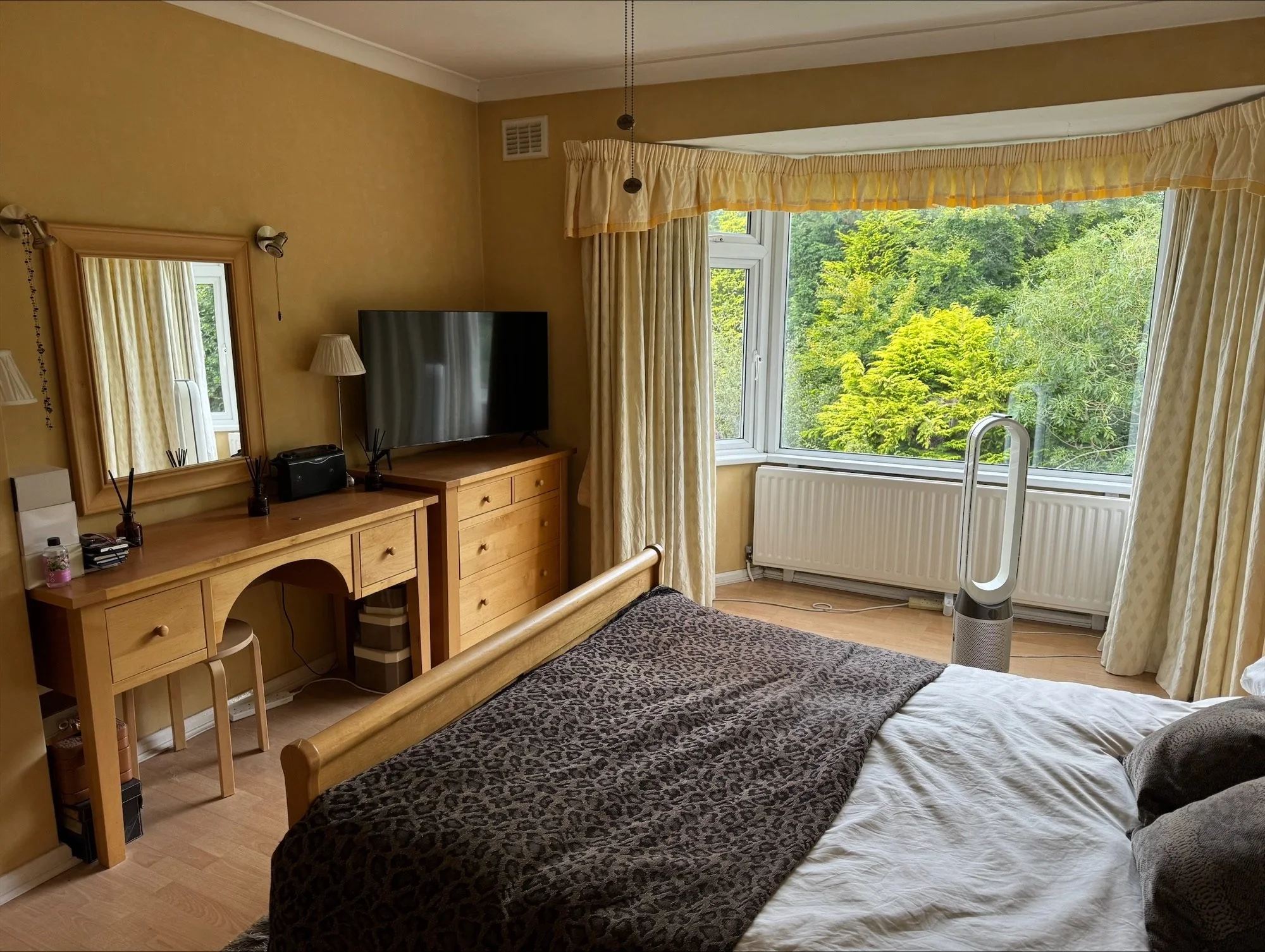 A bedroom with a bed in the foreground covered with a leopard-print blanket. A window showing green trees outside. A TV on a wooden dresser next to a mirror, with a small desk and chair beneath it. A Dyson bladeless fan near the window, with cream-co