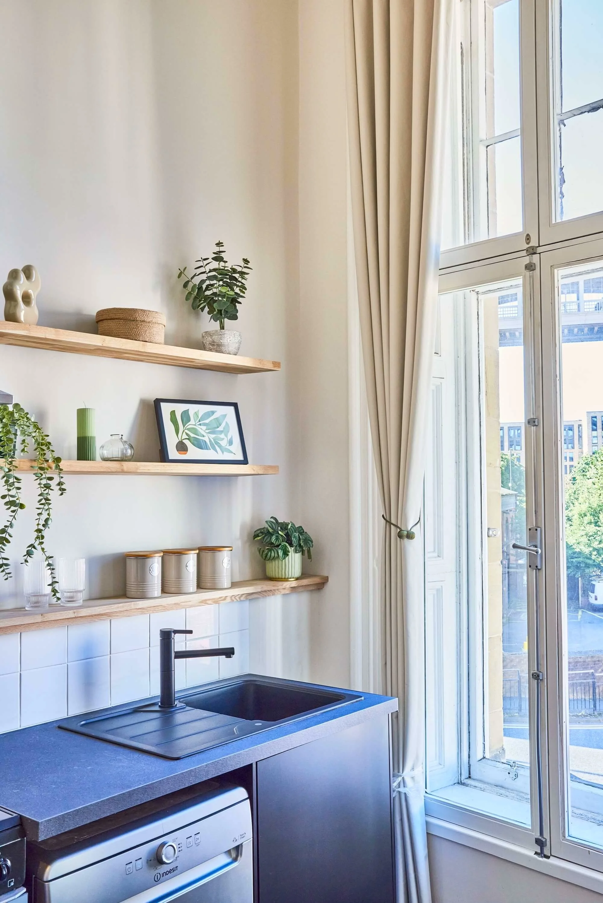 A small kitchen area with a black sink, a black faucet, and a dishwasher below a wooden countertop. Two wooden shelves hold plants, decorative jars, and a framed botanical print. An open window with cream-colored curtains lets in natural light, revea